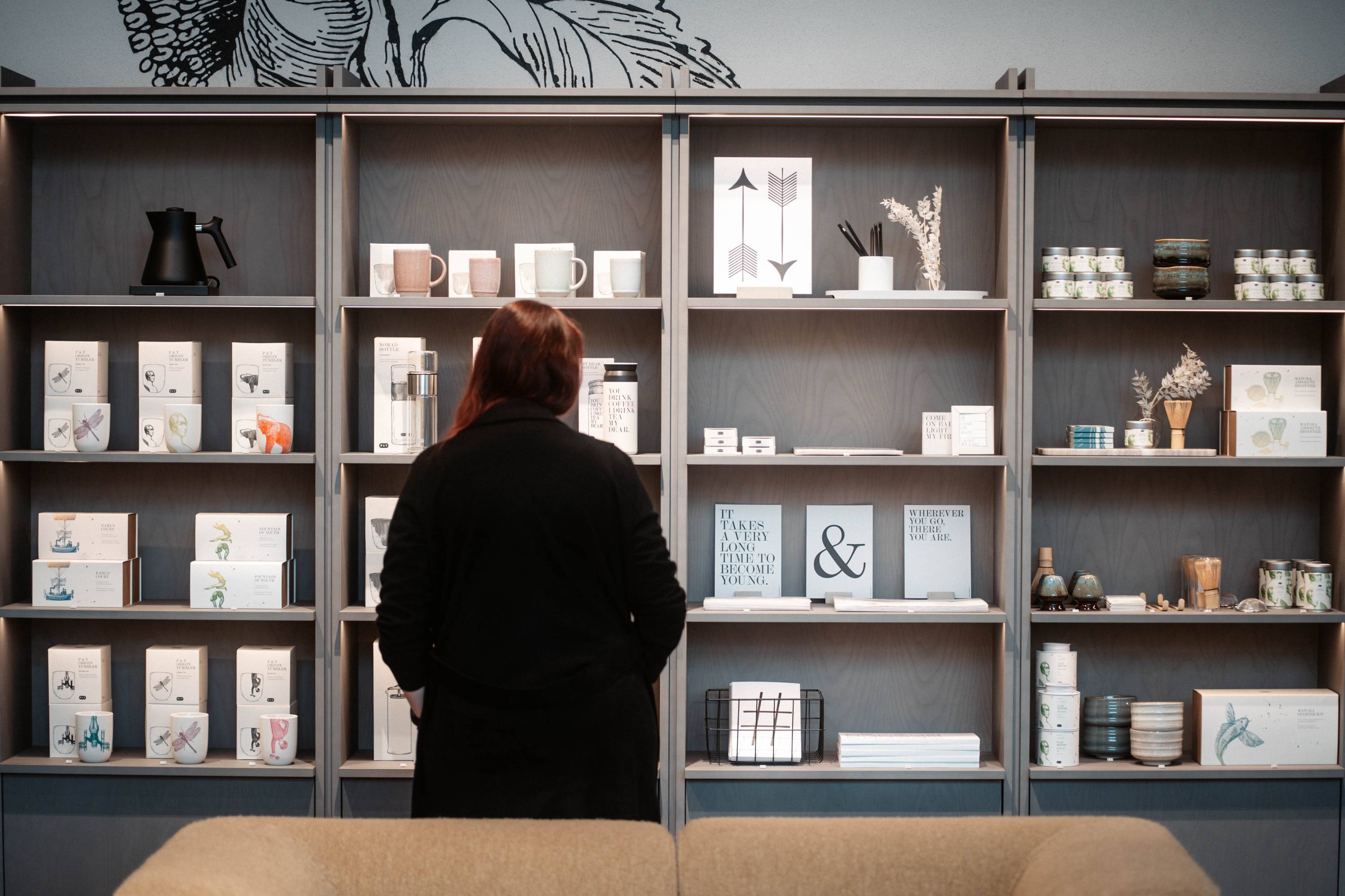 Person im schwarzen Mantel betrachtet Regale mit Teebechern in ruhiger Umgebung