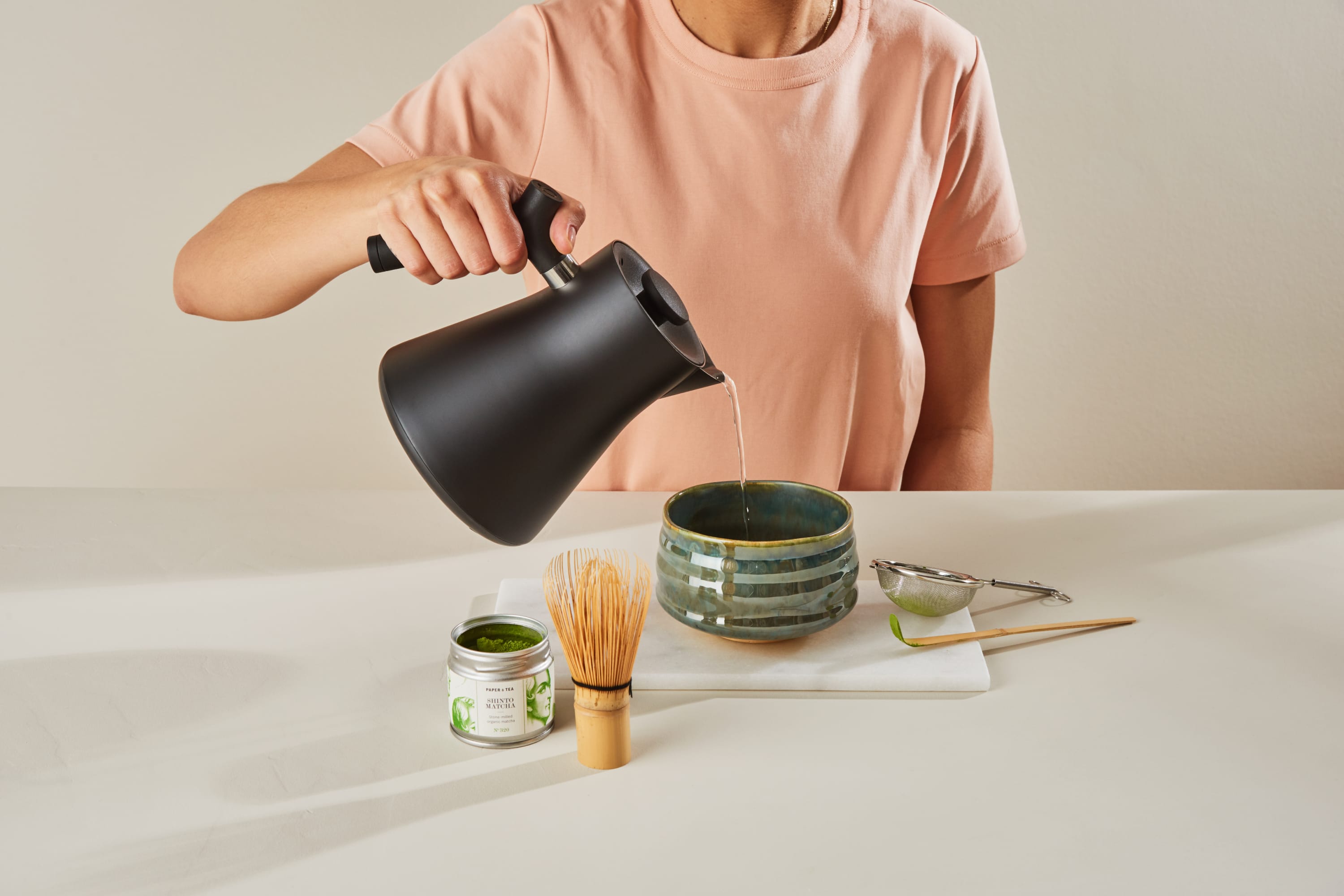 Person in rosa Shirt gießt heißes Wasser aus schwarzem Wasserkocher in grüne Keramikschale, auf dem Tisch Matcha-Zubehör: offenes Matcha-Glas, Bambusbesen, Metallsieb mit Matcha, weißes Tuch und Bambuslöffel