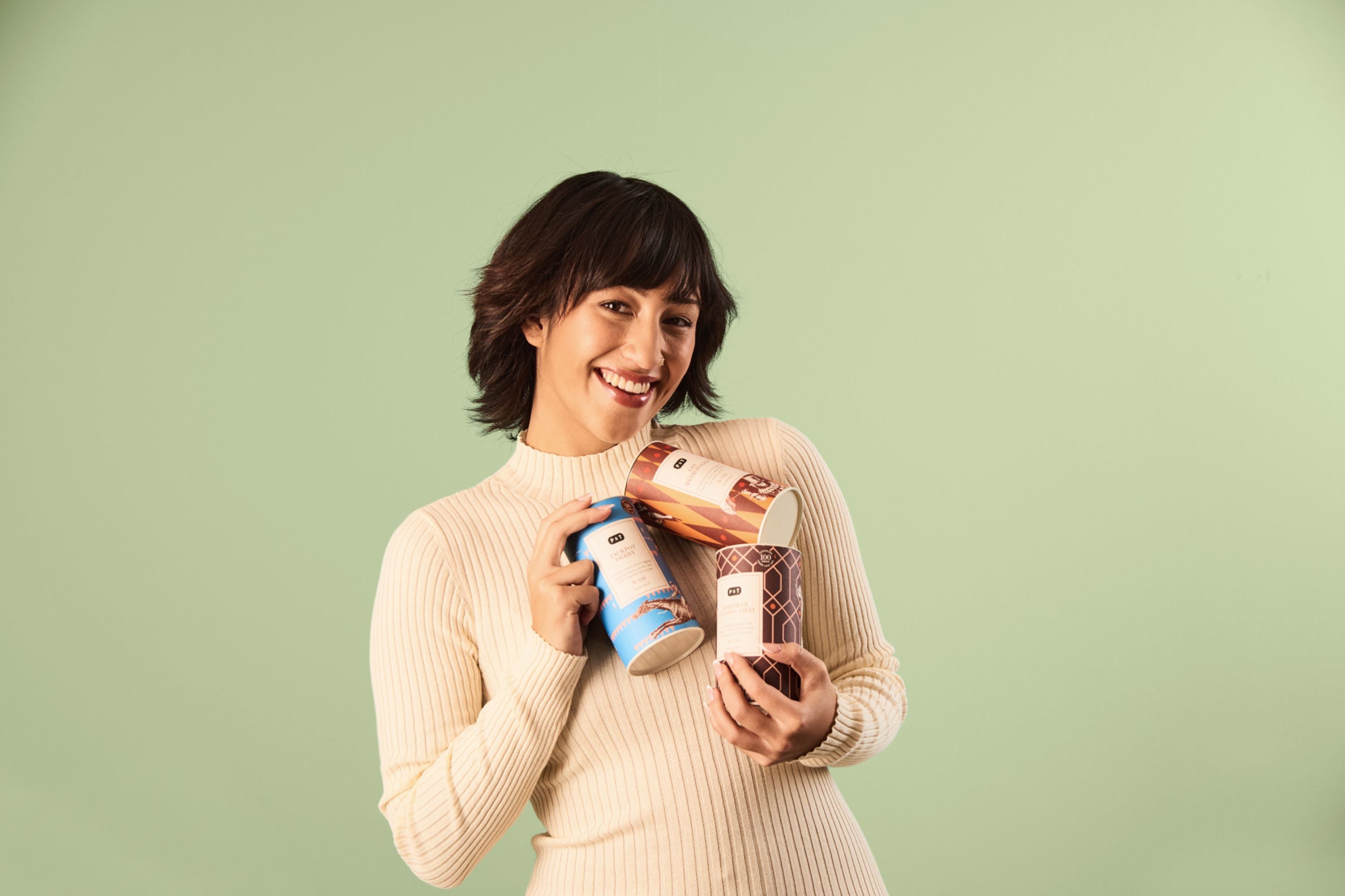 Person in hellem, geripptem Pullover hält drei Teedosen vor einem hellgrünen Hintergrund