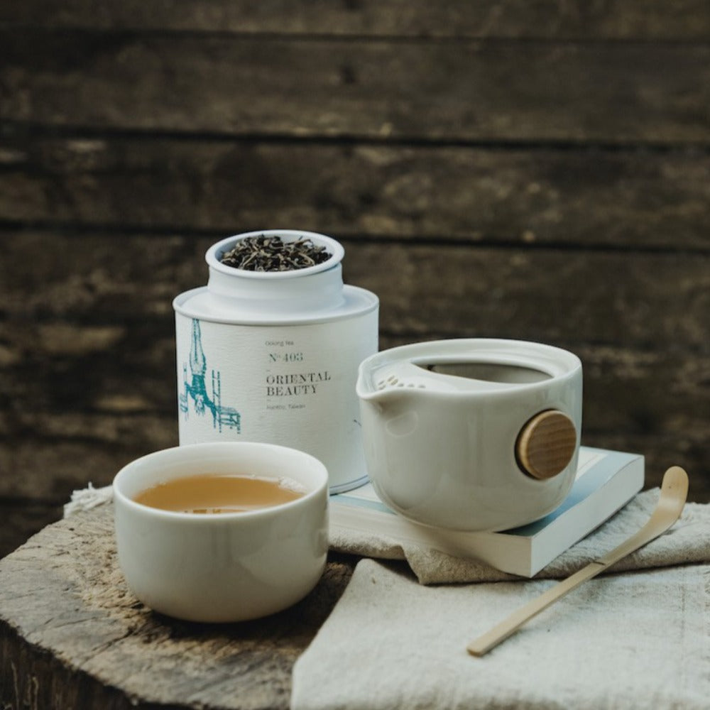 Weißes Keramikgefäß mit losem Oriental Beauty Tee, helle Teetasse und Teekanne auf rustikalem Holz.