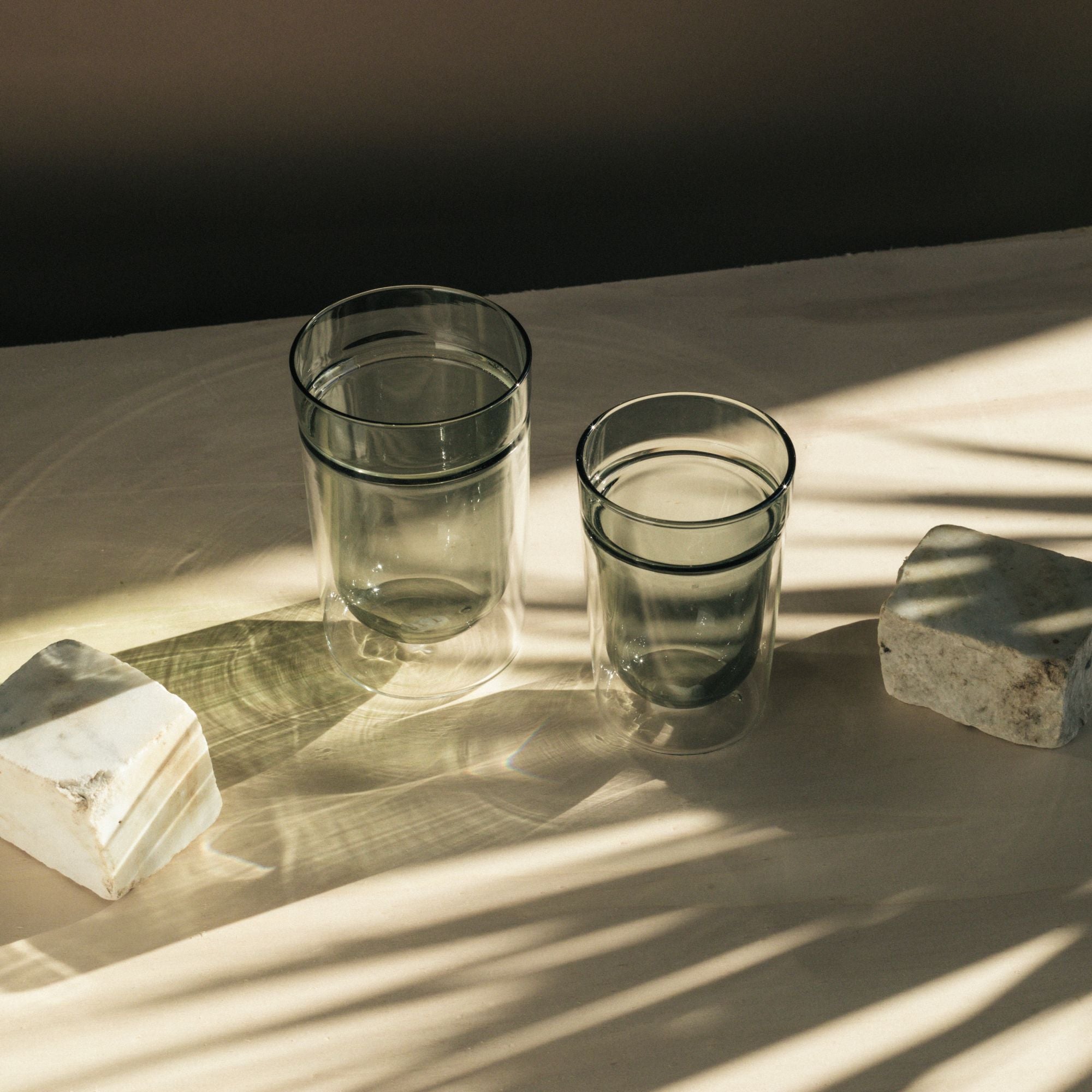 Zwei doppelwandige Gläser auf hellem Hintergrund, eines halb mit Wasser gefüllt, neben unregelmäßigen Steinen.