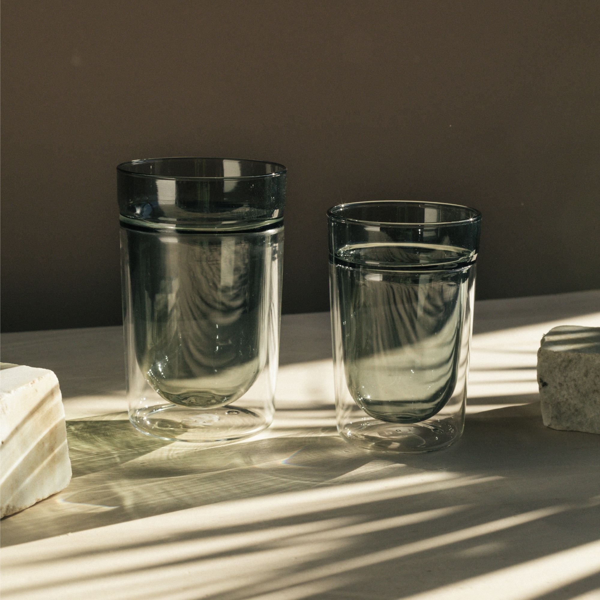 Zwei doppeltwandige Gläser mit Wasser auf heller Oberfläche, umgeben von weißen Steinen und sanften Lichtmustern.