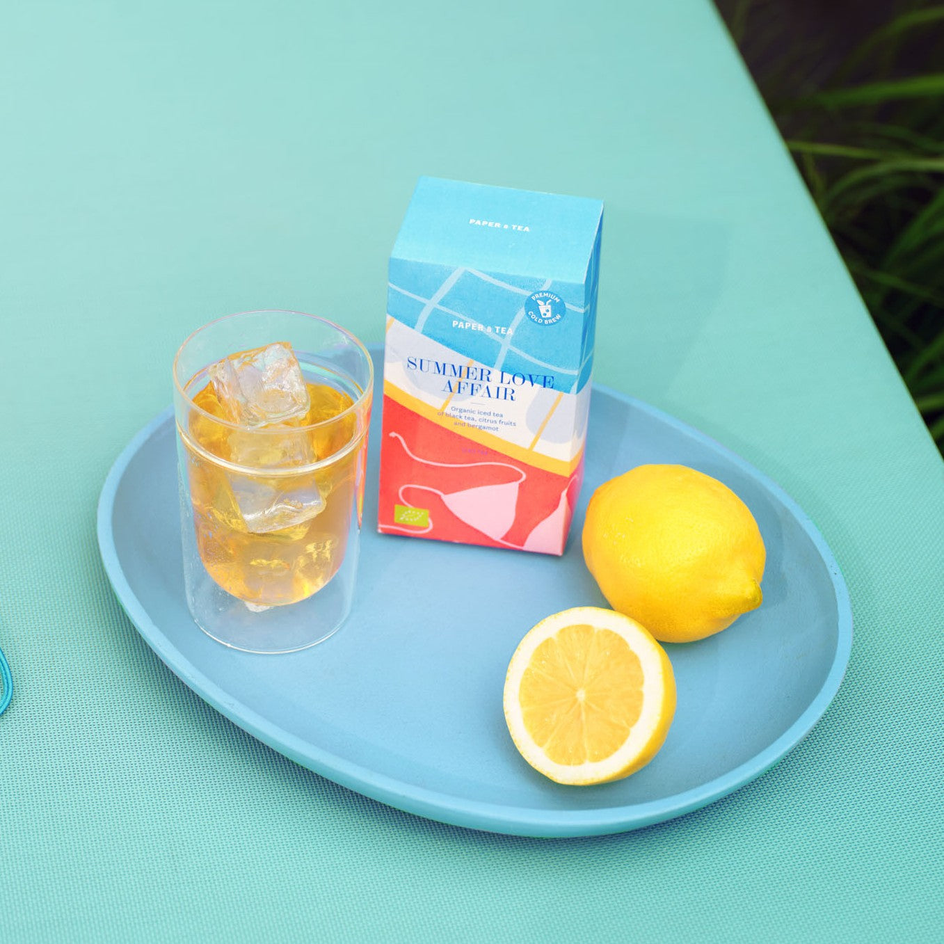 Bunter BIO-Eisteekarton von PAPER & TEA mit schwarzem Tee, Zitrus und Bergamotte auf blauem Tablett.