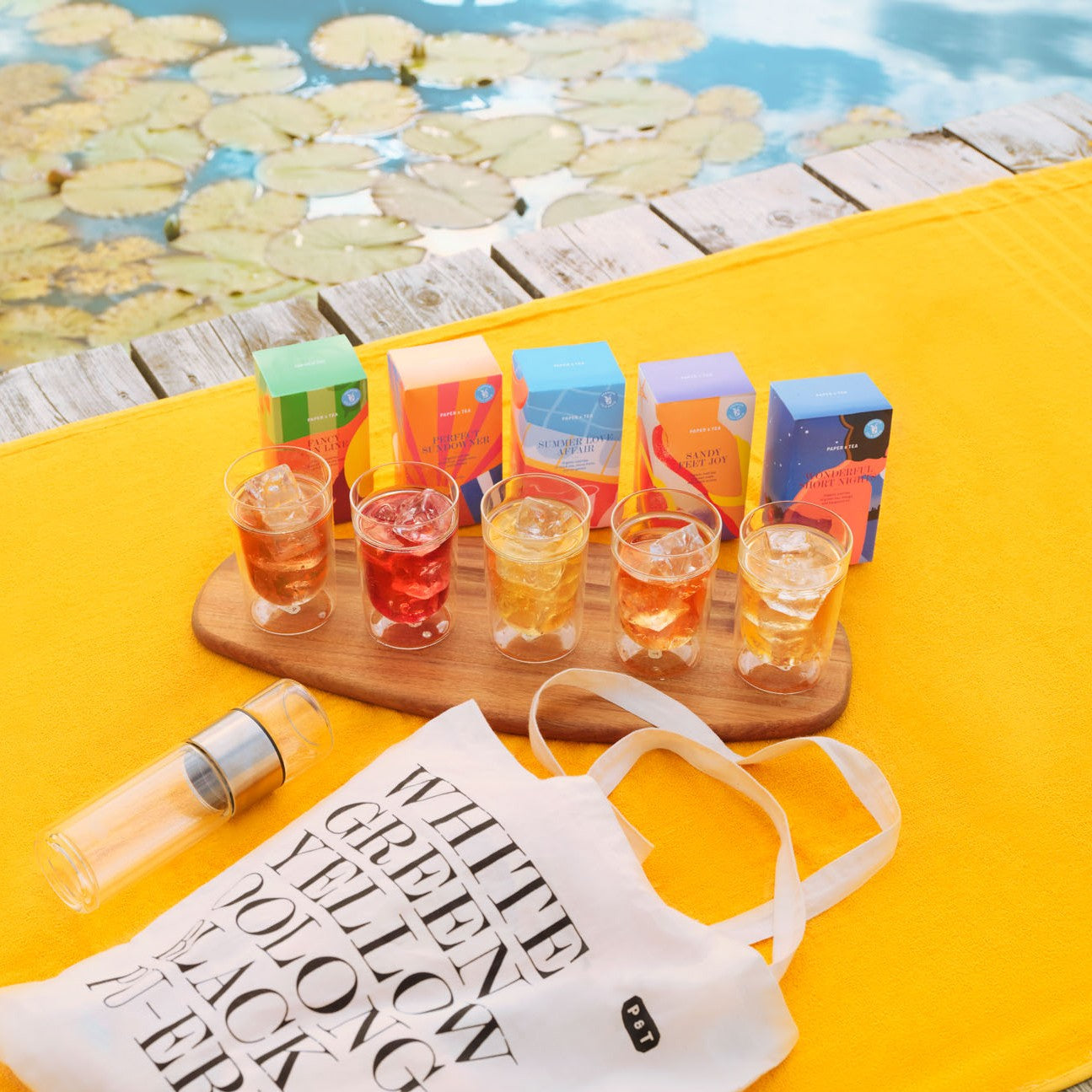 Fünf bunte TEA & JOY Teeschachteln mit Gläsern Eistee auf Holztablett am Steg mit Seerosen und Tasche