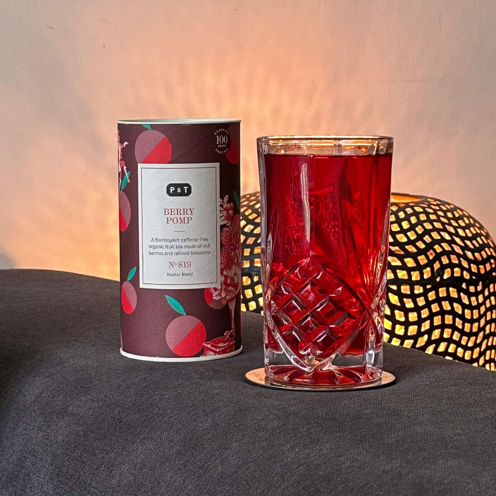 Beerenmuster-Dose von P & T BERRY POMP neben Glas mit rotem Früchtetee vor warm leuchtender Lampe.