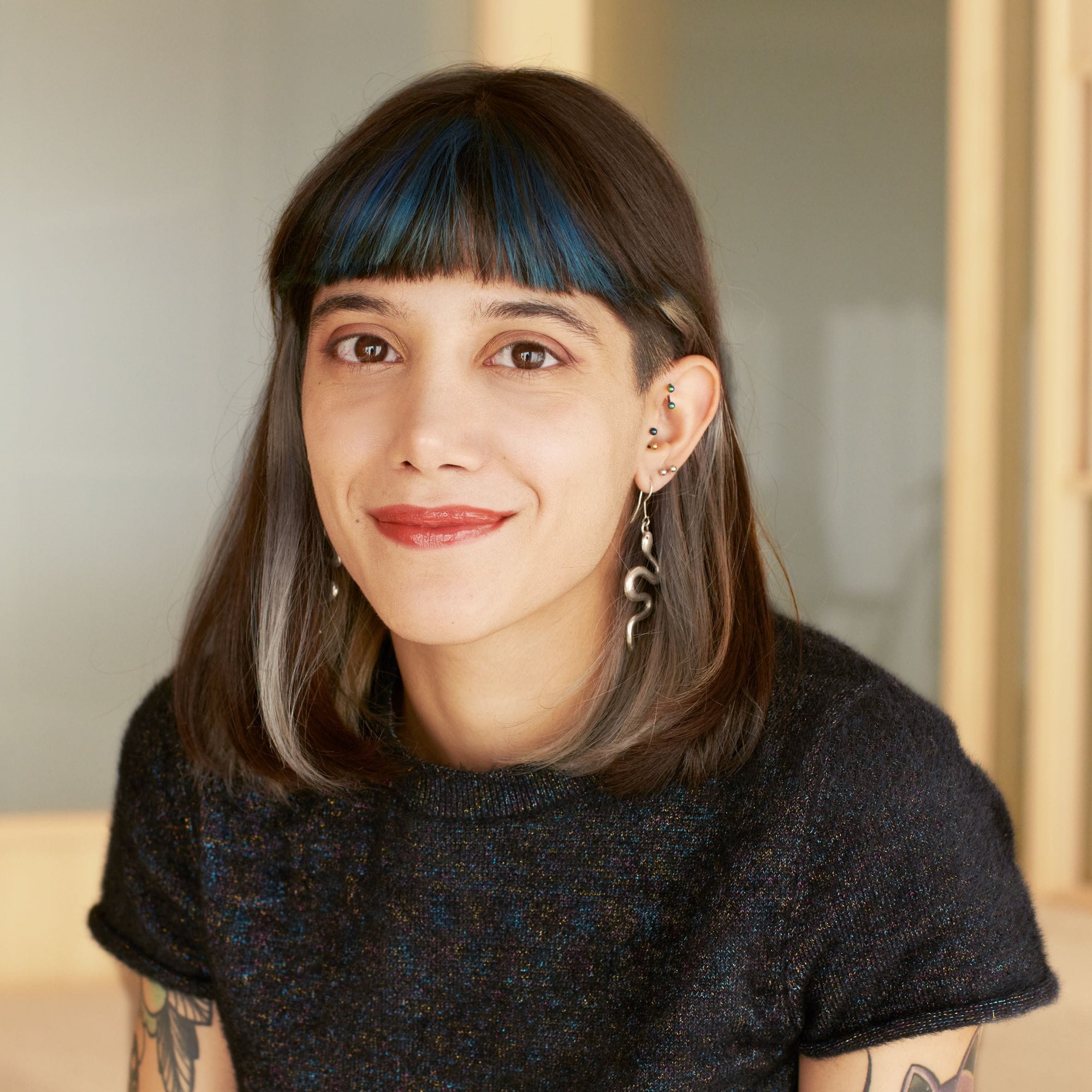 Junge Person mit blauen Haarsträhnen, gesprenkeltem Pullover und Tattoos im warmen Innenraum.