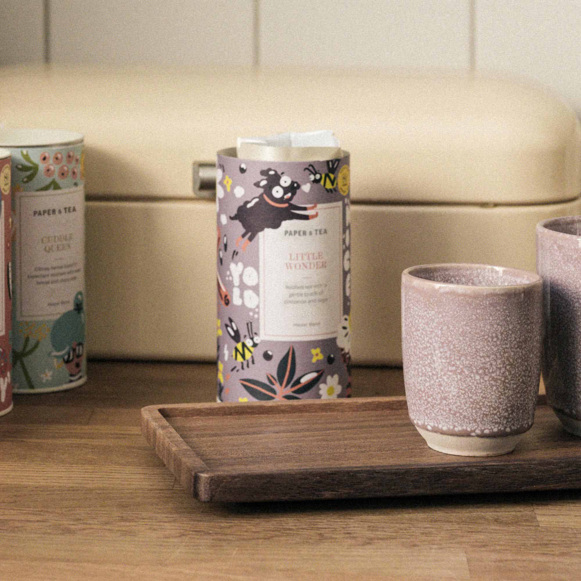 Lila Zylinderbehälter PAPER & TEA LITTLE WONDER mit Bienen und Hund, violette Tassen auf Holztablett und bunte Teedosen.