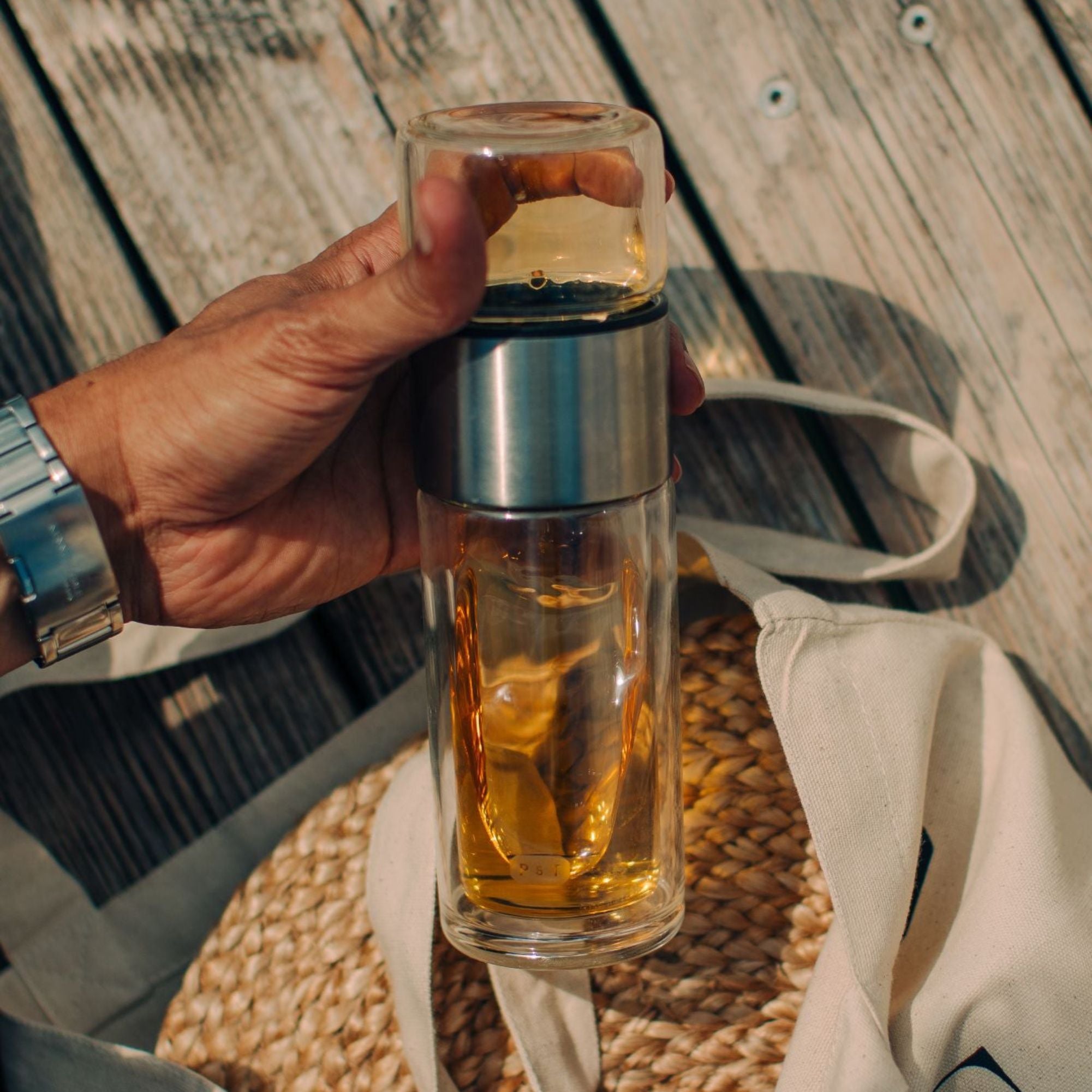 Hand mit silberner Uhr hält doppelwandige Glasflasche mit Tee über Stofftasche auf Naturfasermatte.
