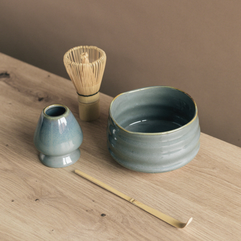 Teeset mit blauer Keramikschüssel, Bambusbesen und Löffel auf hellem Holztisch vor beiger Wand.