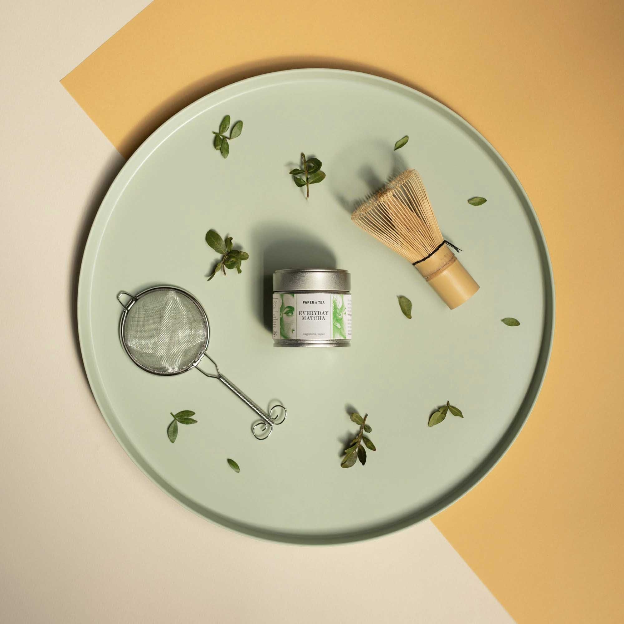 Hellgrünes Tablett mit Matcha-Dose, Bambus-Whisk, Teesieb und frischen Teeblättern.