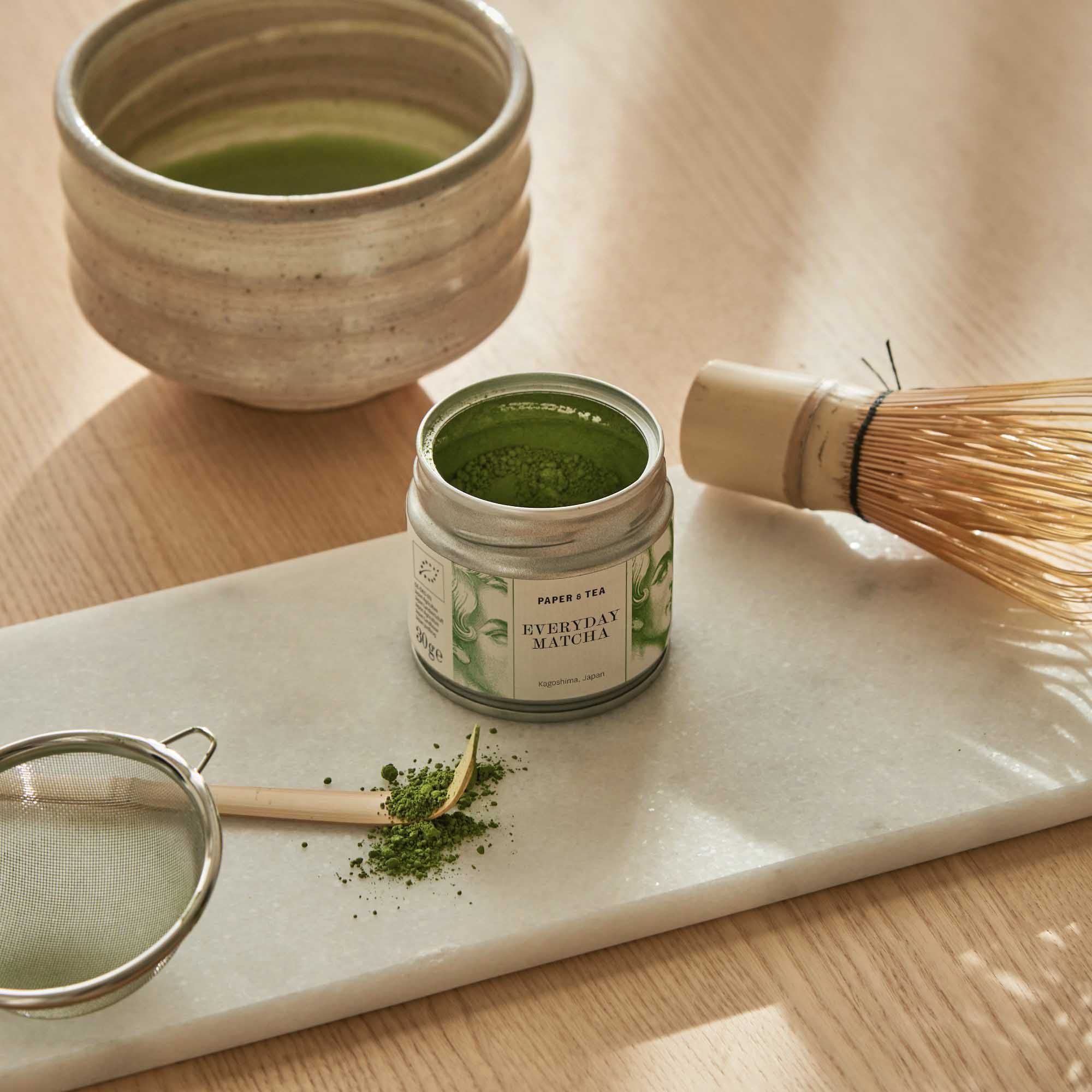 Dose mit grünem Matcha-Pulver, Bambusbesen, Teesieb und Keramiktasse auf Holztisch