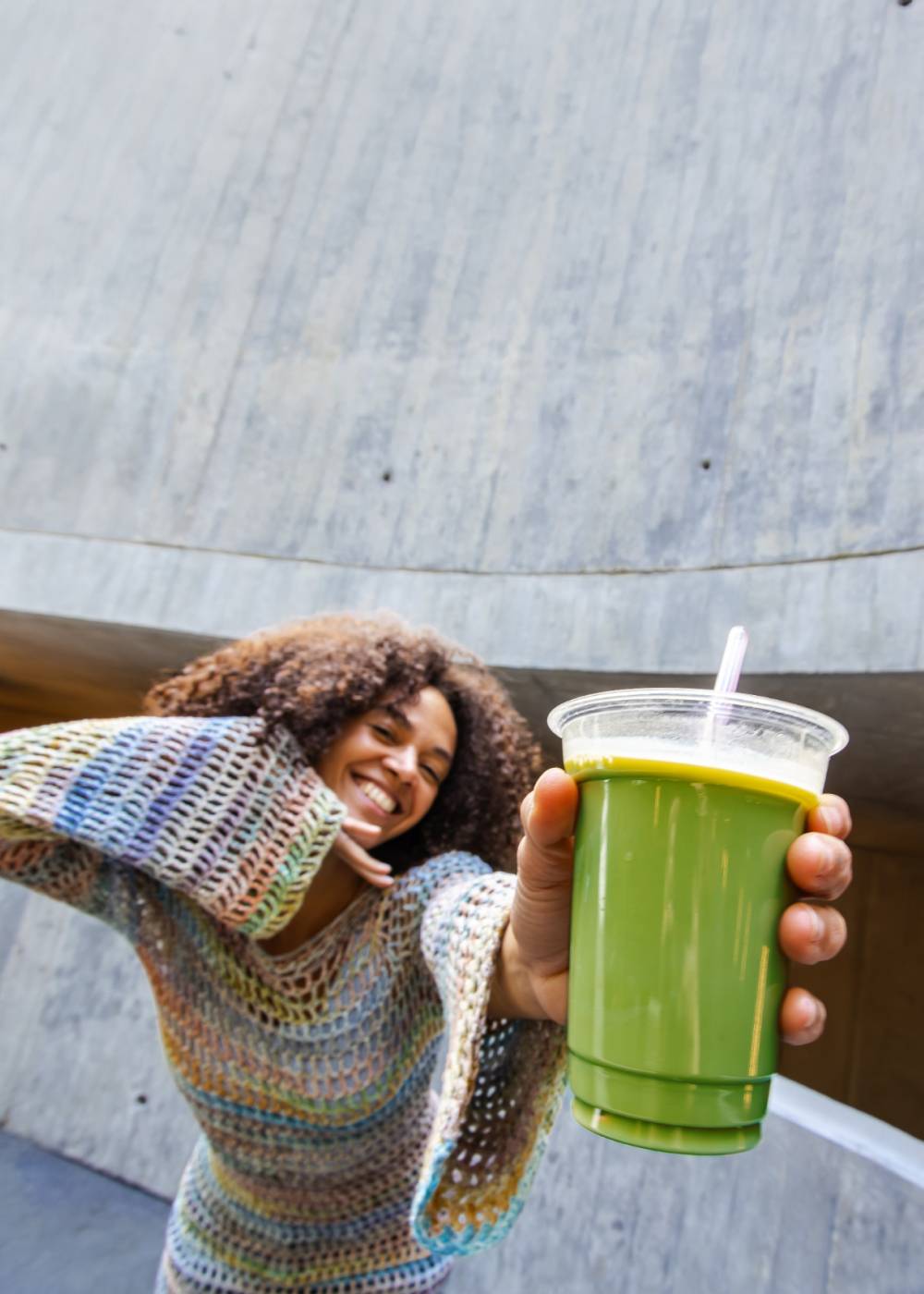 Junge Frau im bunten Häkelpullover mit grünem Smoothie im Becher vor urbanem Betonhintergrund.