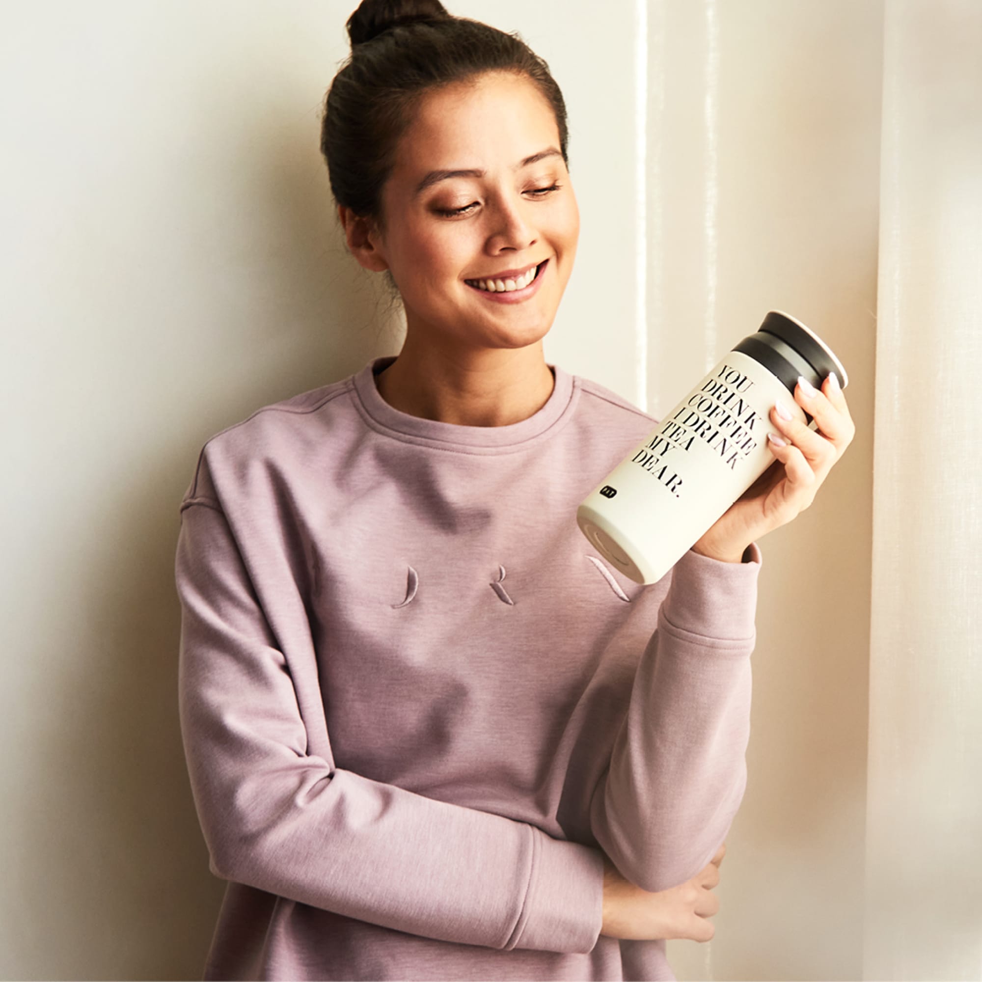 Junge Frau im lila Sweatshirt hält weißen Becher mit 'YOU DRINK COFFEE I DRINK TEA MY DEAR' vor hellem Hintergrund.