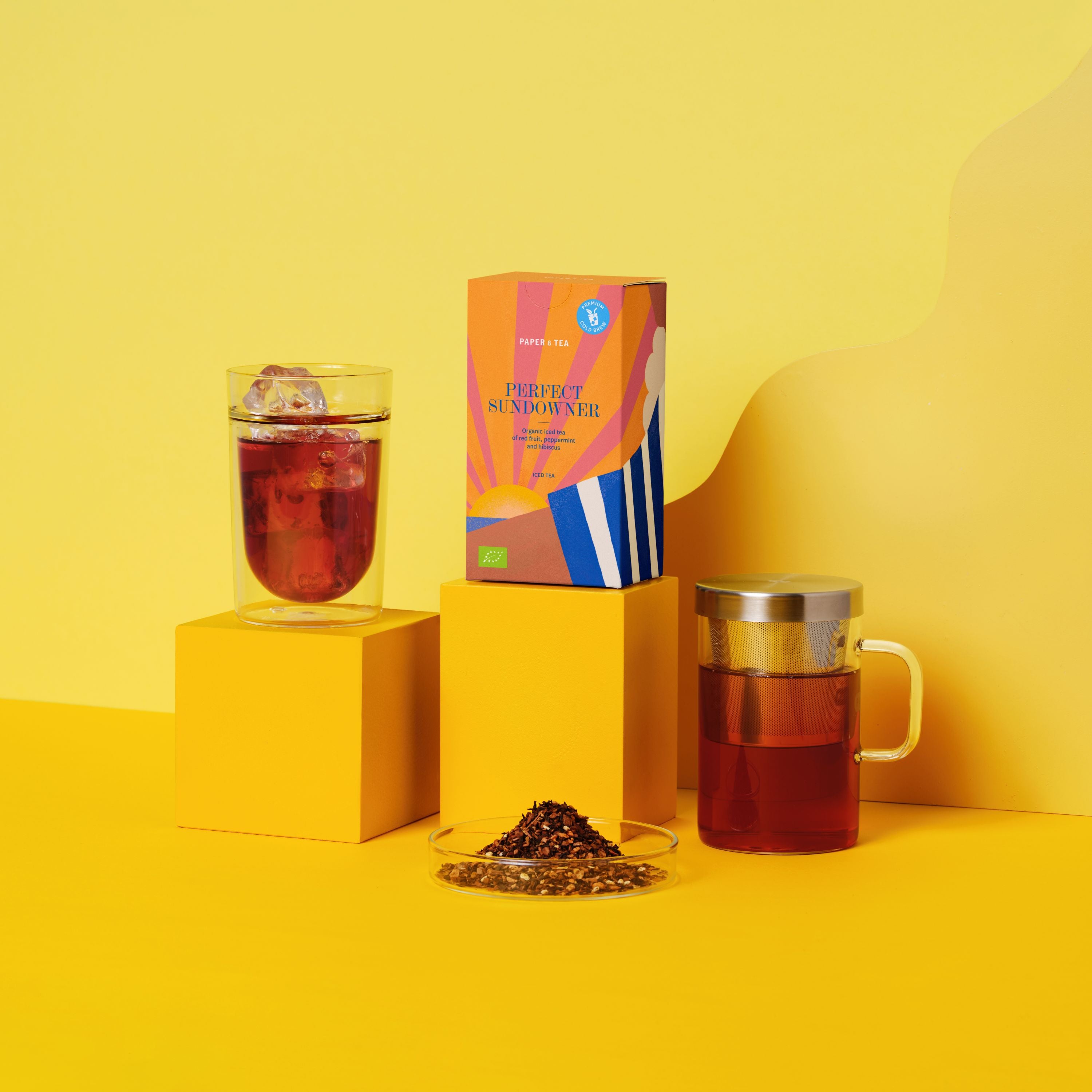Bunte Paper & Tea Teeverpackung 'Perfect Sundowner' mit Sonnenmotiv, Glas mit Eis und Teekanne vor gelbem Hintergrund.