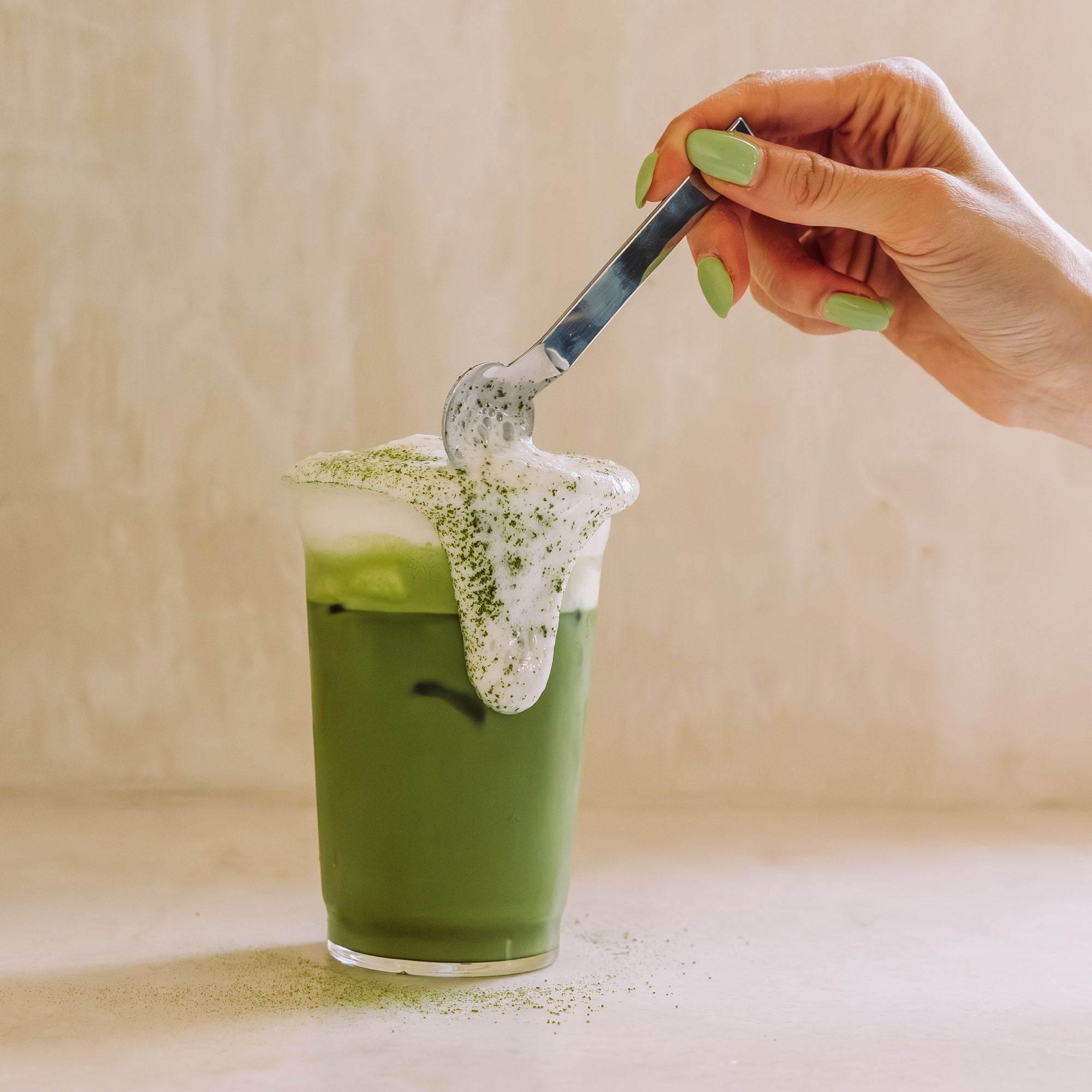 Klares Glas mit grünem Eistee und schaumiger Matcha-Schicht, Hand mit grünen Nägeln schöpft Schaum mit Löffel.