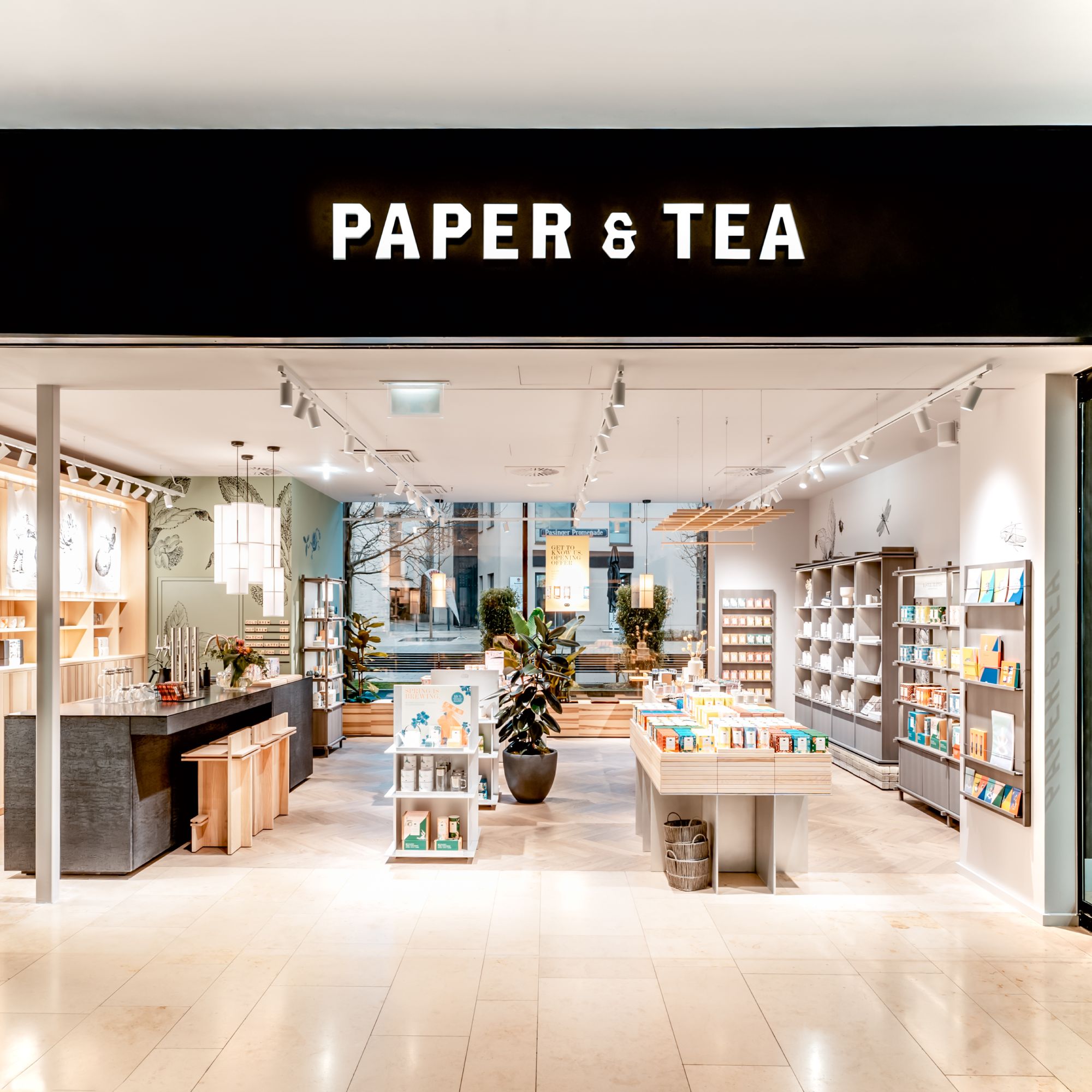 Moderne PAPER & TEA Verkaufsfiliale mit Holzfußboden, Regalen voller Teeprodukte und großen Fenstern zur Straße.