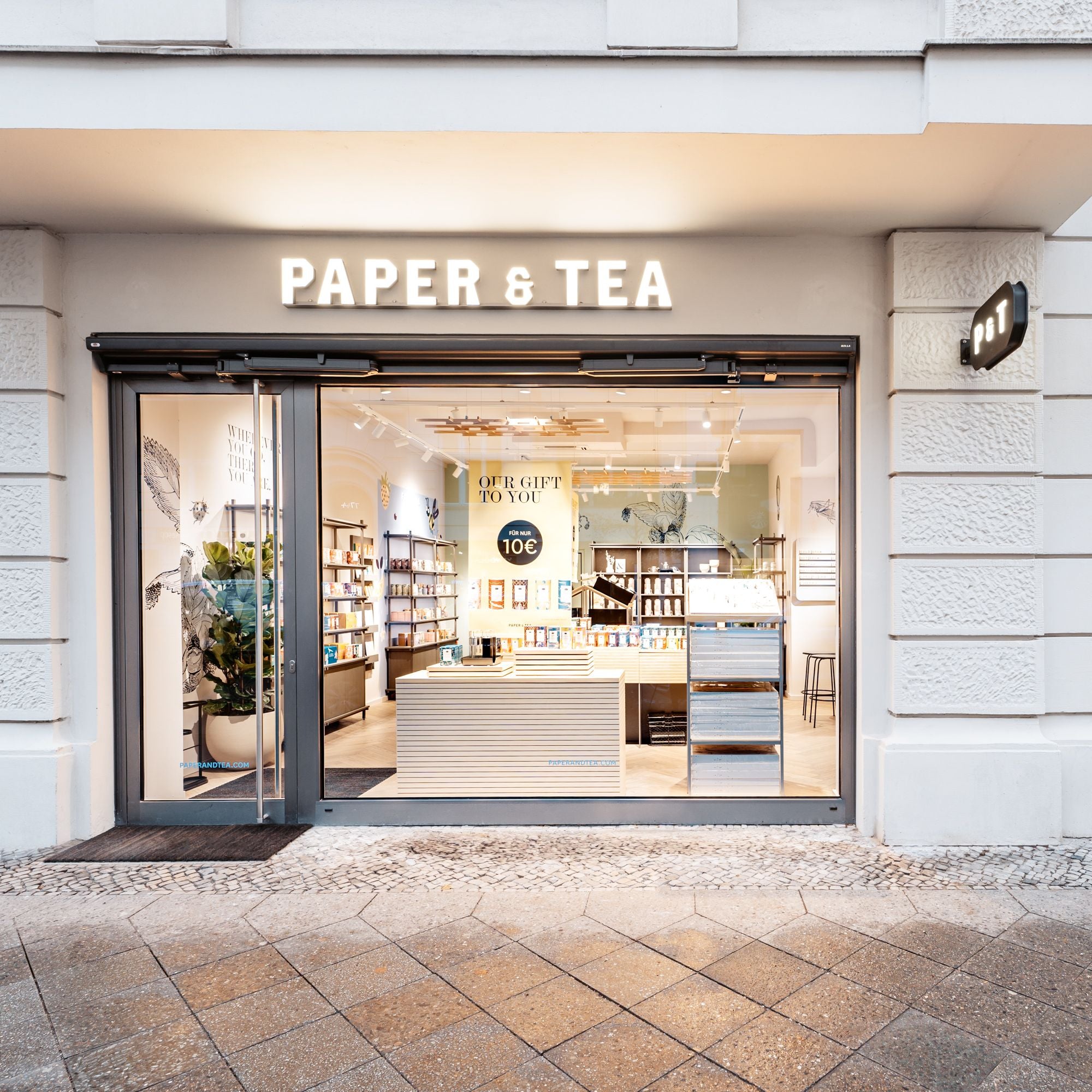 Modernes PAPER & TEA Ladengeschäft mit gläserner Fassade, Holztresen und 10-Euro-Angebotsschildern drinnen.
