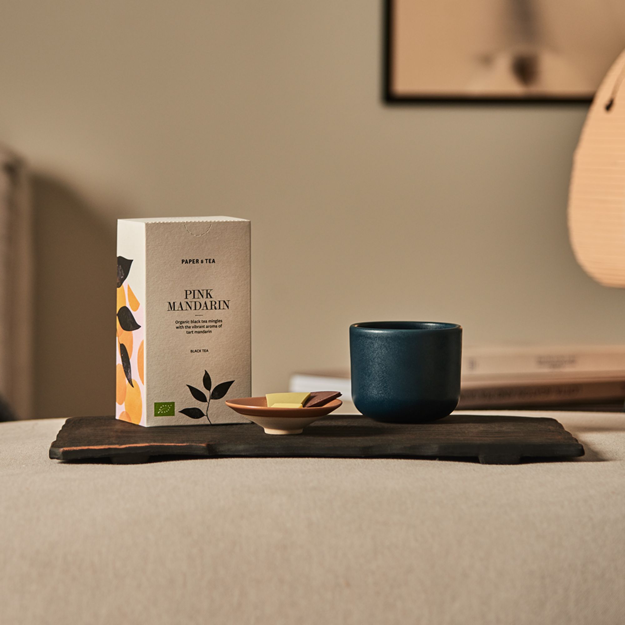 Box mit PAPER & TEA PINK MANDARIN Bio-Schwarztee auf dunklem Holzteller, neben Keramikbecher und Schale mit Schokolade.