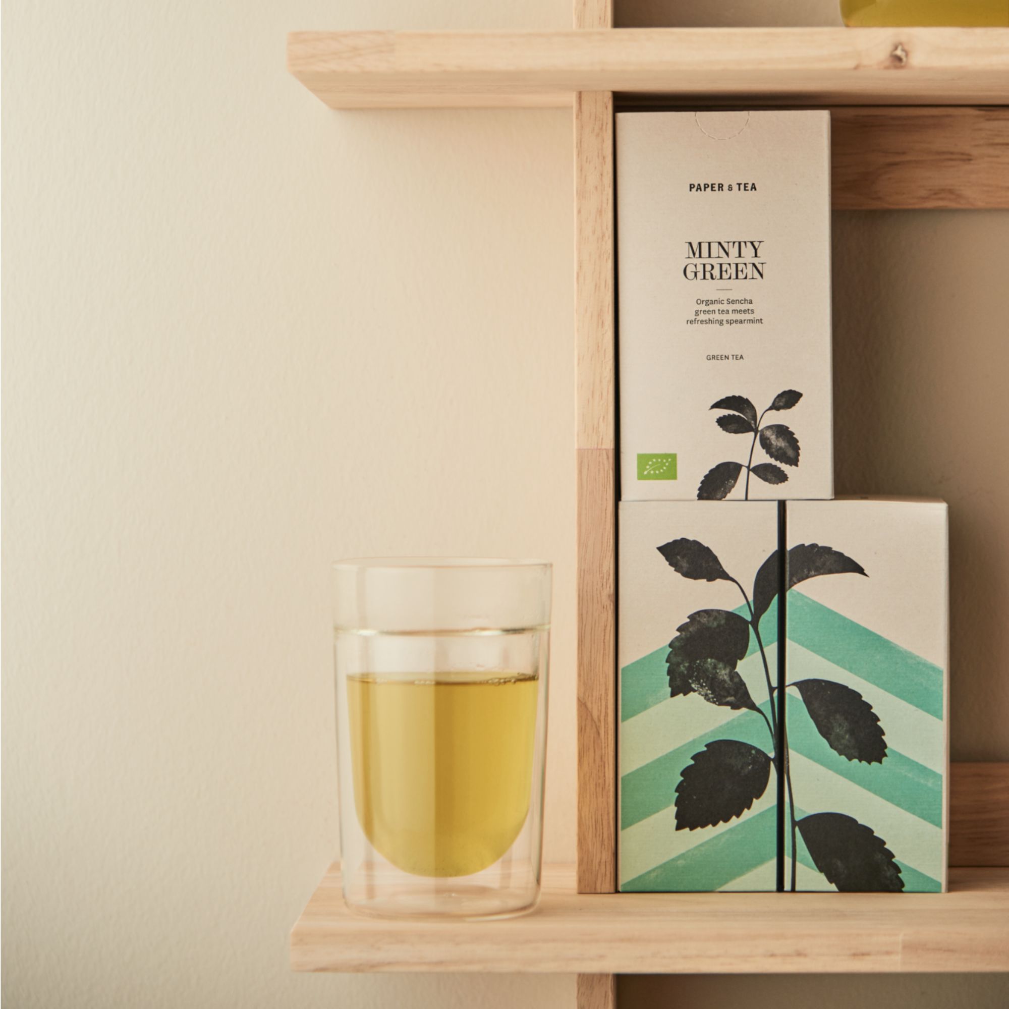 Beige PAPER & TEA-Verpackung mit "Minty Green" neben Glas Sencha Grüntee auf Holzregal.