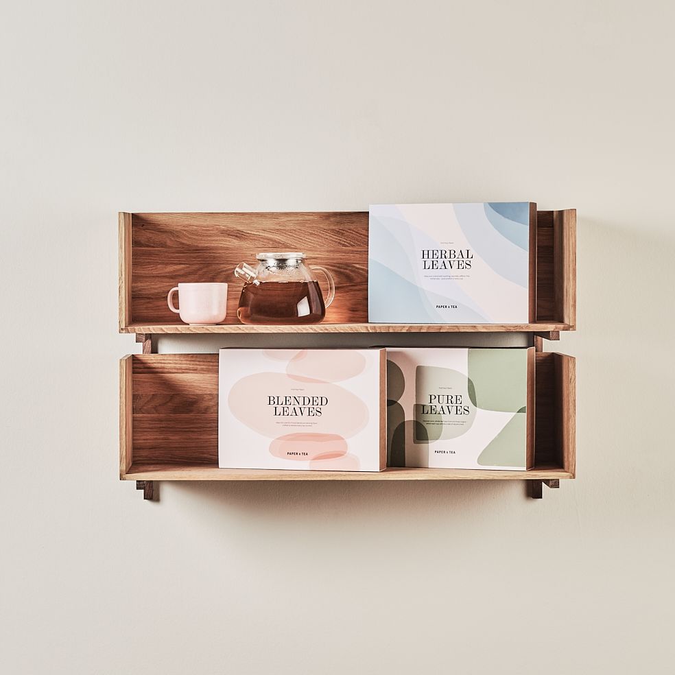 Minimalistisches hölzernes Wandregal mit drei Teeboxen, Glas-Teekanne mit dunklem Tee und rosa Keramikbecher.