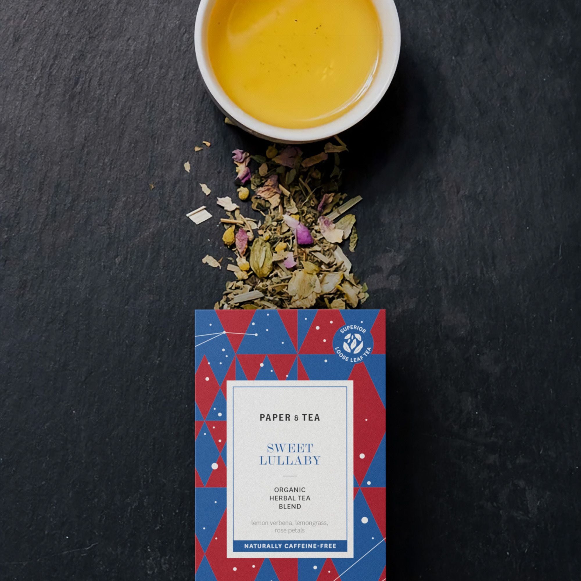 Draufsicht auf bunte Paper & Tea Sweet Lullaby Teepackung und goldener Tee in Tasse auf dunklem Hintergrund.