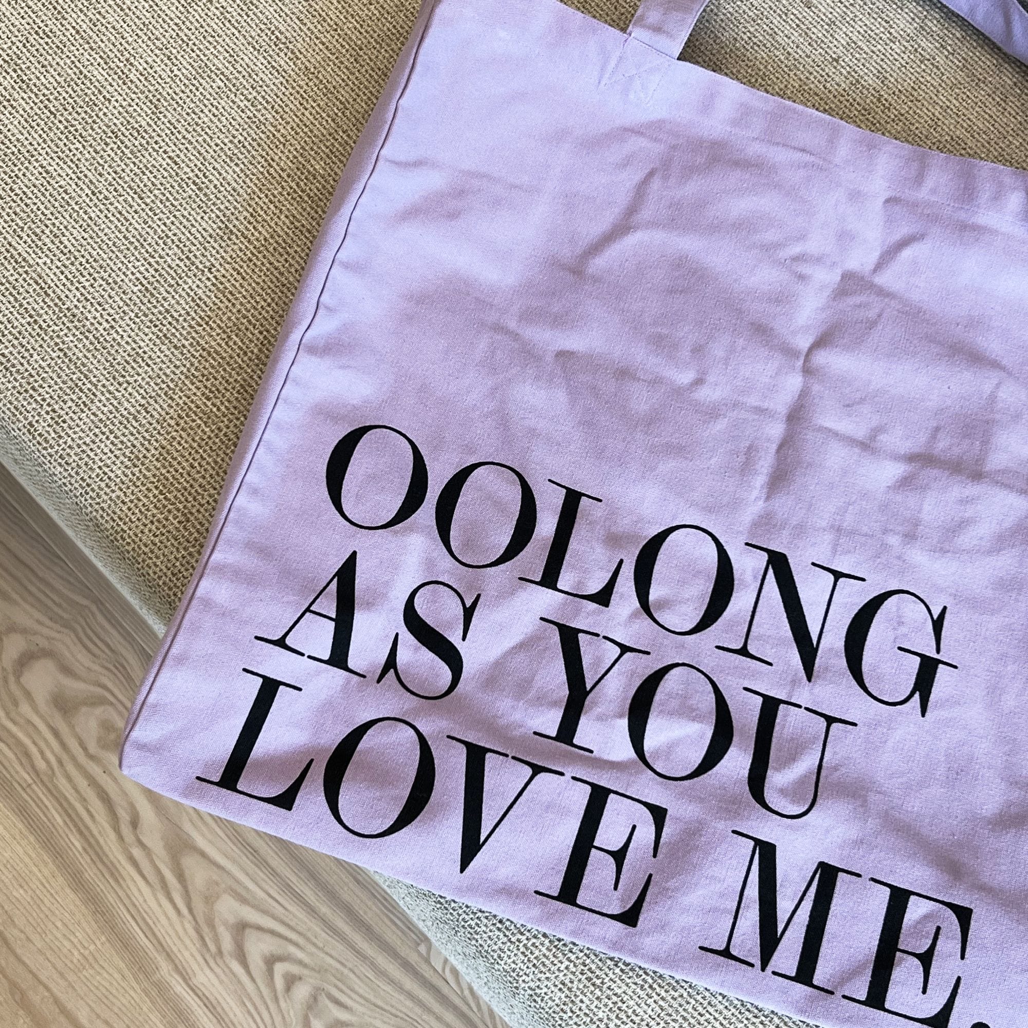 Lavendelfarbene Baumwolltasche mit schwarzem Aufdruck 'OOLONG AS YOU LOVE ME', auf Holzboden