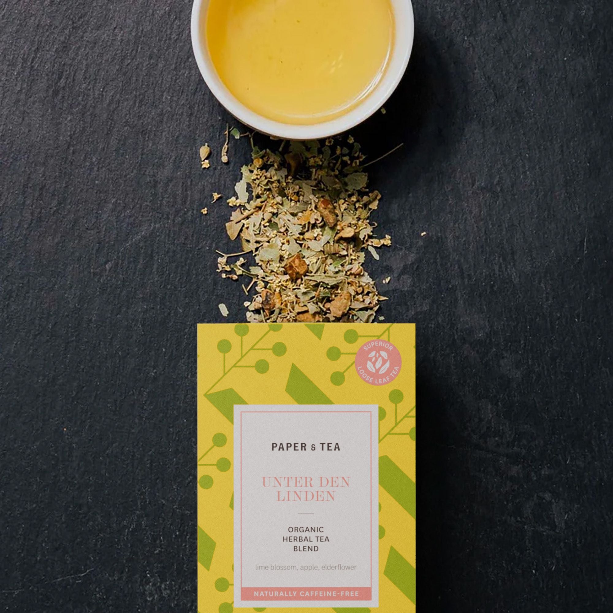 Tasse mit hellem Kräutertee neben losem Lindenblütentee und gelber Tee-Verpackung.
