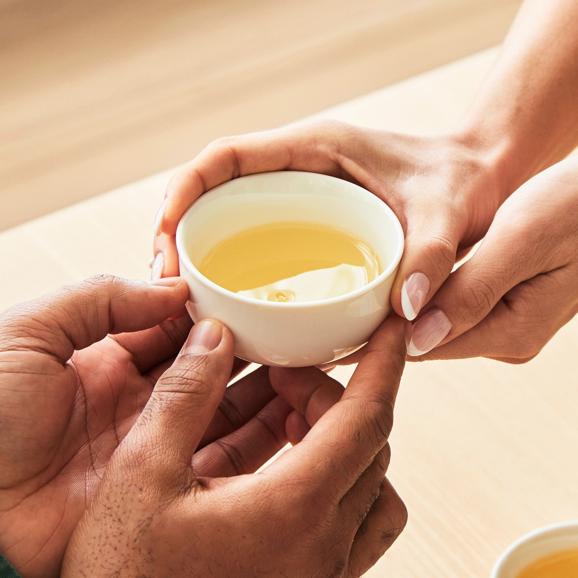 Zwei Paar Hände unterschiedlicher Hauttöne halten eine weiße Tasse mit hellem Tee über einem Holztisch.