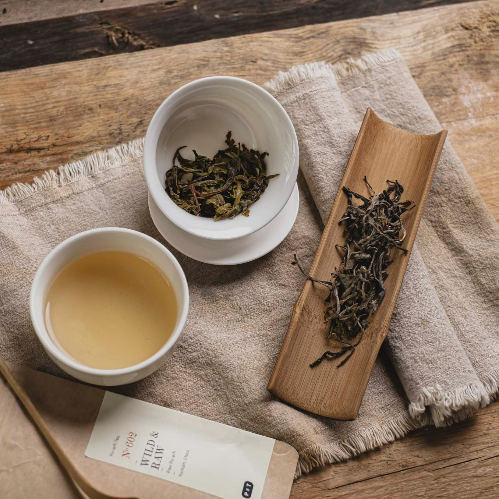 Weiße Keramiktasse mit hellem Pu-erh Tee, Gaiwan mit nassen Teeblättern, lose Roh-Pu-erh-Blätter auf Holztablett, No 602 Packung.