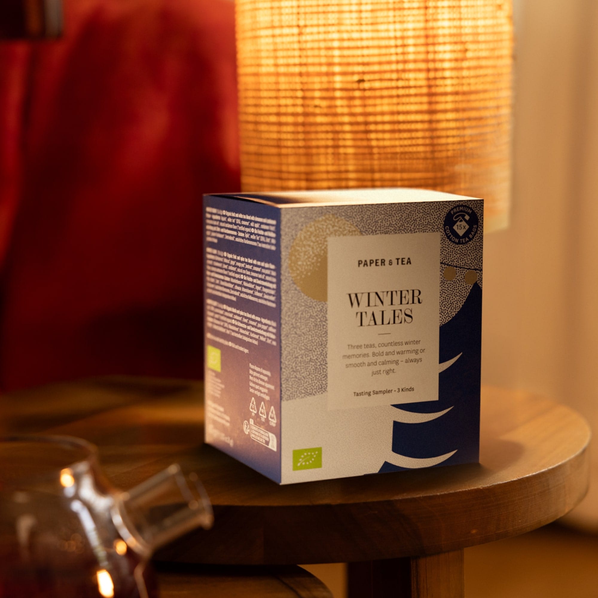 Winter Tales Tee Probierbox von PAPER & TEA auf Holztisch mit sanftem Lampenlicht und Teekanne im Hintergrund.