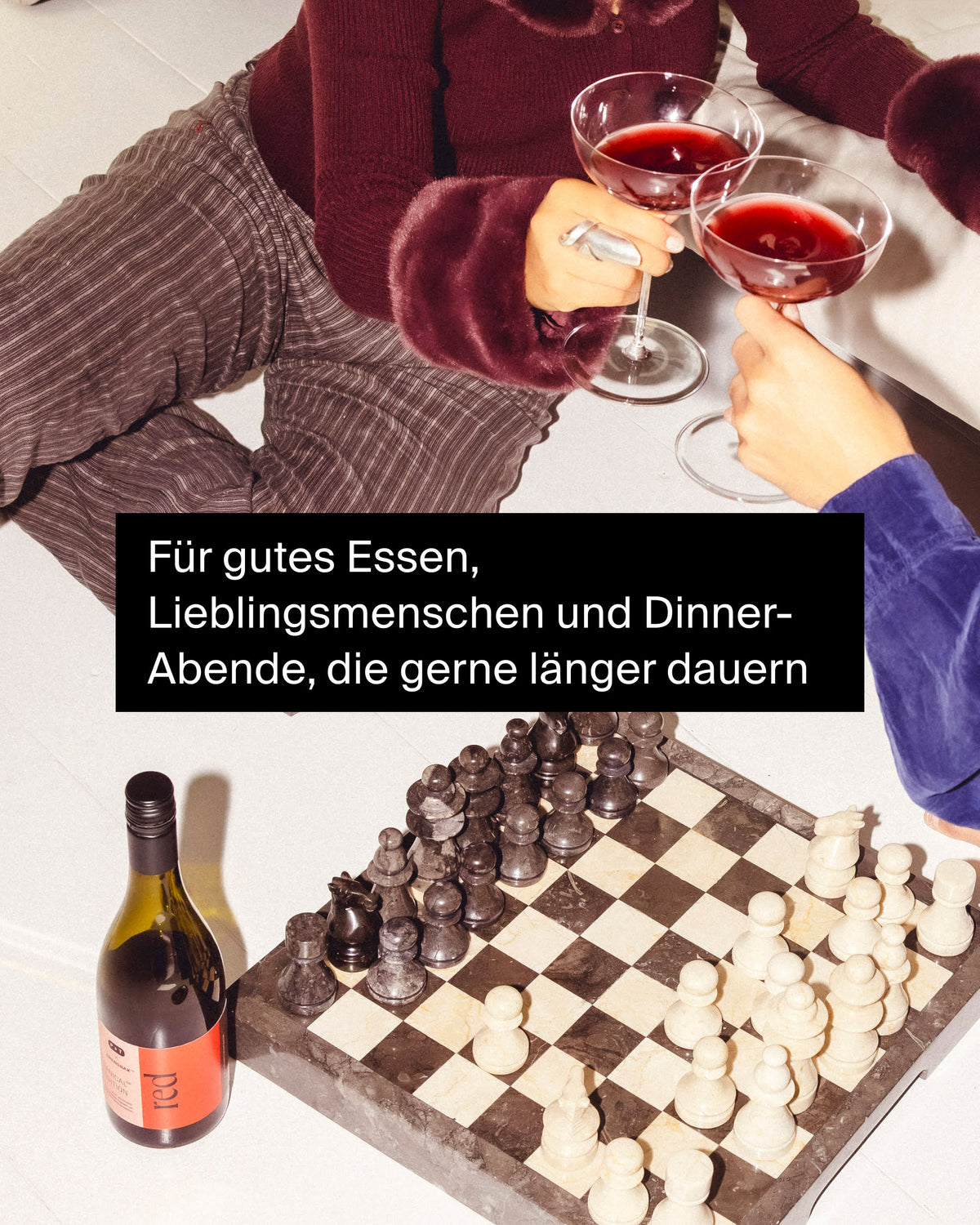 Zwei Personen stoßen mit roten Cocktails an, vor einem Marmorschachbrett und Weinflasche in gemütlicher Abendstimmung.