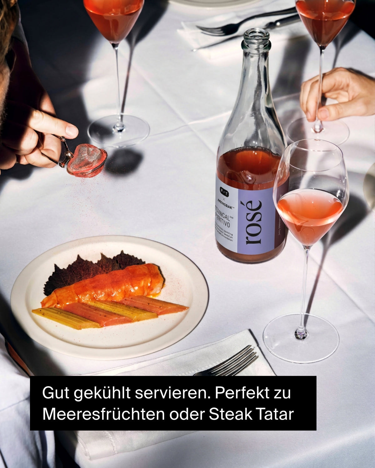 Flasche Roséwein mit lila Etikett, drei Gläser Rosé und glasiertes Meeresfrüchtegericht auf weißem Tisch
