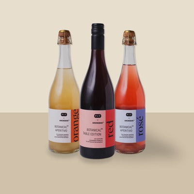 Drei Arkenstone Botanical Aperitivo Flaschen in Orange, Rot und Rosé auf beigem Hintergrund.