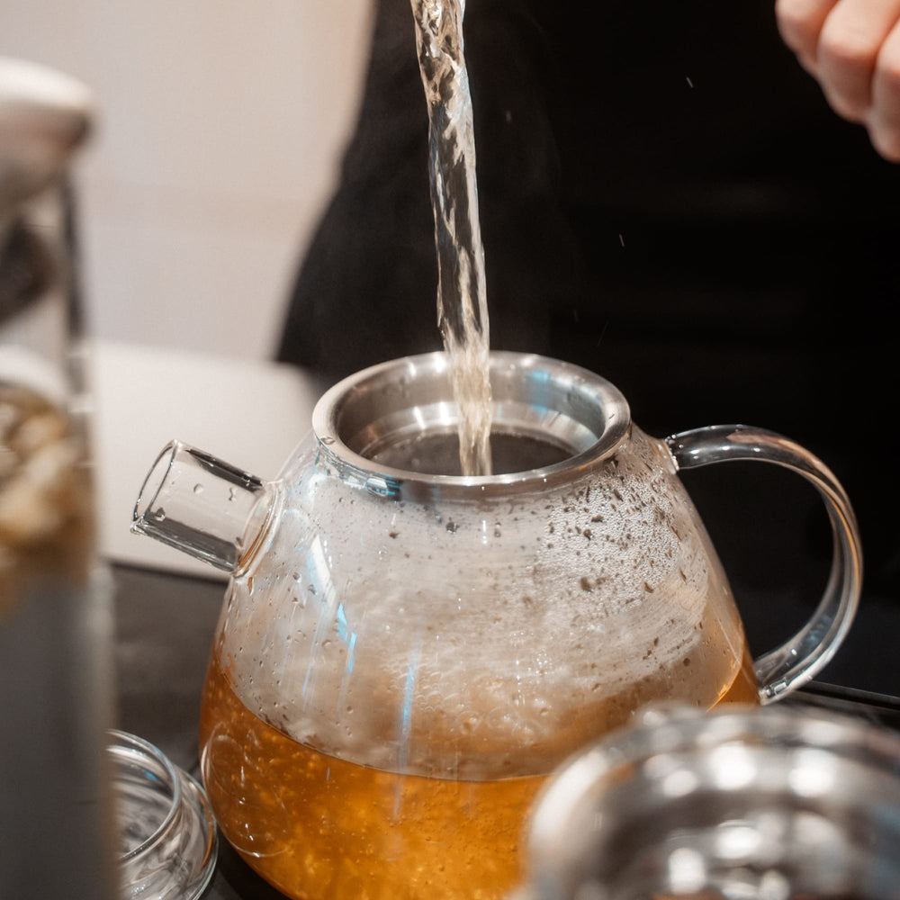 Glas-Teekessel mit Metallrand füllt sich mit heißem, bernsteinfarbenem Tee. Kondensation und Dampf sichtbar.