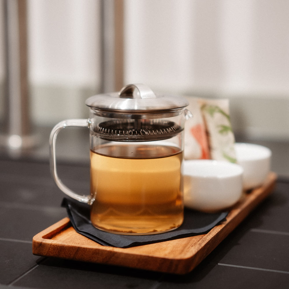 Transparente Glas-French-Press mit hellem Bernsteintee auf Holztablett, zwei weiße Keramiktassen, Teebeutel mit Blattmuster.