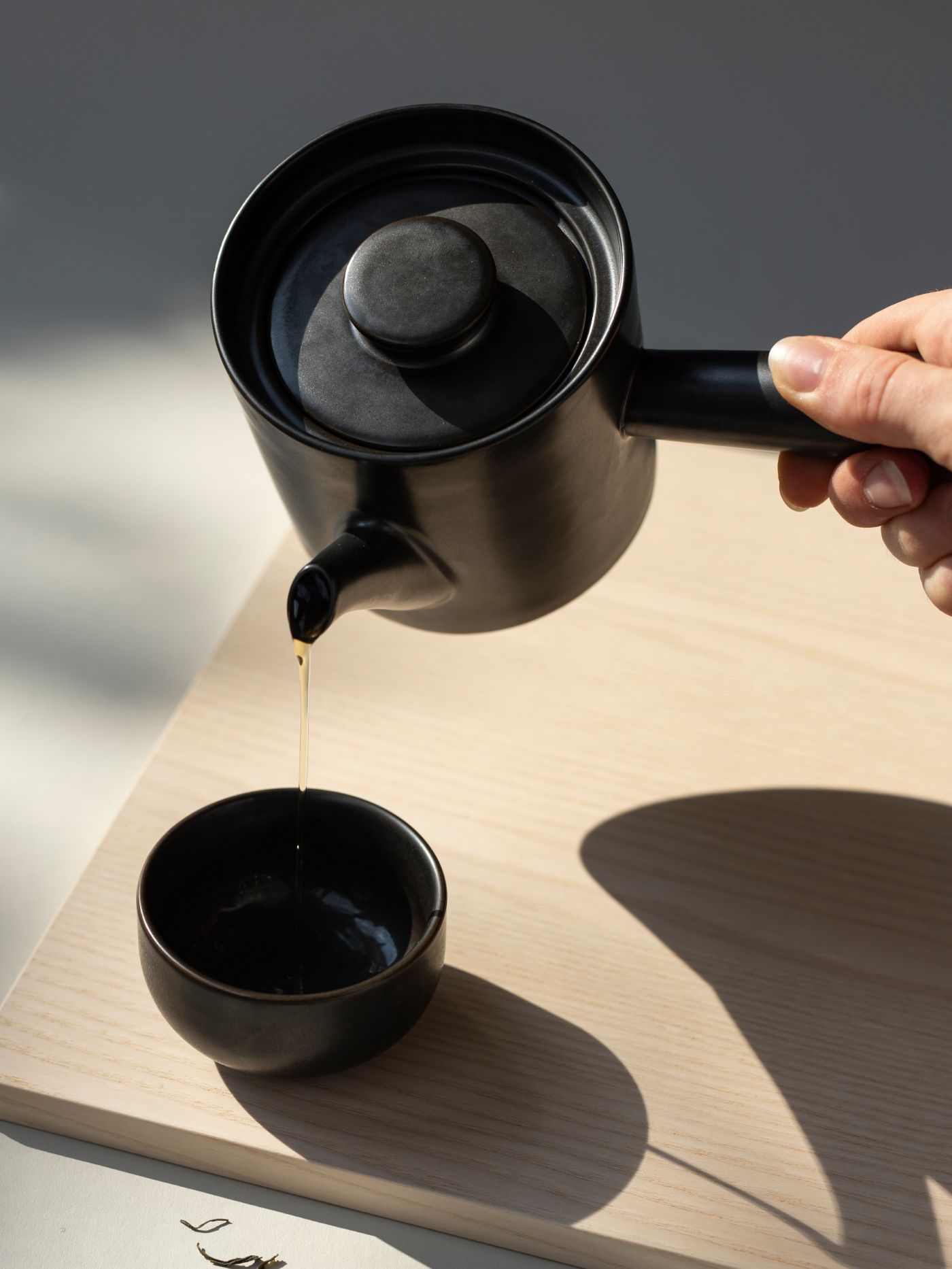 Schwarze Teekanne gießt Tee in kleine Tasse auf hellem Holztisch bei natürlichem Licht