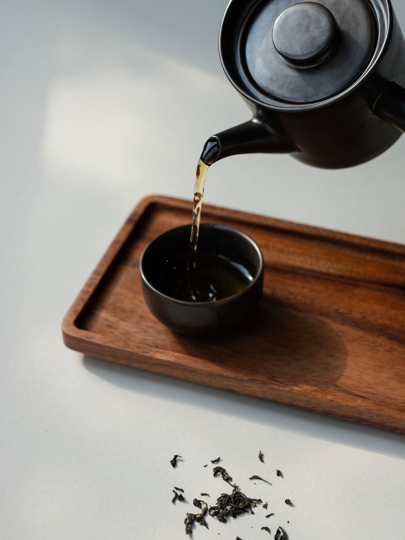 Schwarze Keramik-Teekanne gießt Tee in kleine Tasse auf Holztablett, umgeben von losen Teeblättern.