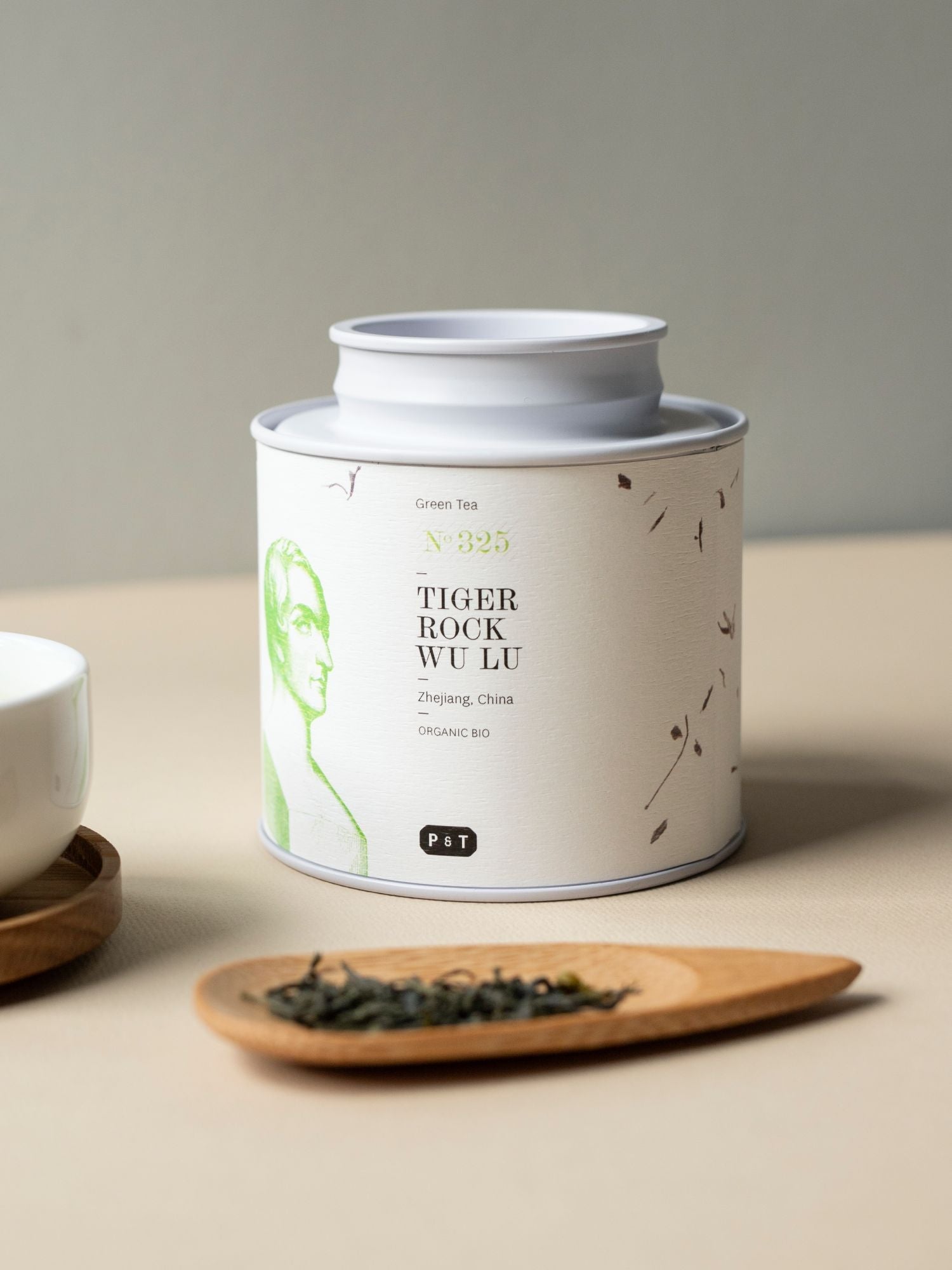 Weißer Teebehälter mit grünem Label ‚BIO-Tiger Rock Wu Lu‘, lose Blätter auf Holzlöffel, neben weißer Tasse.