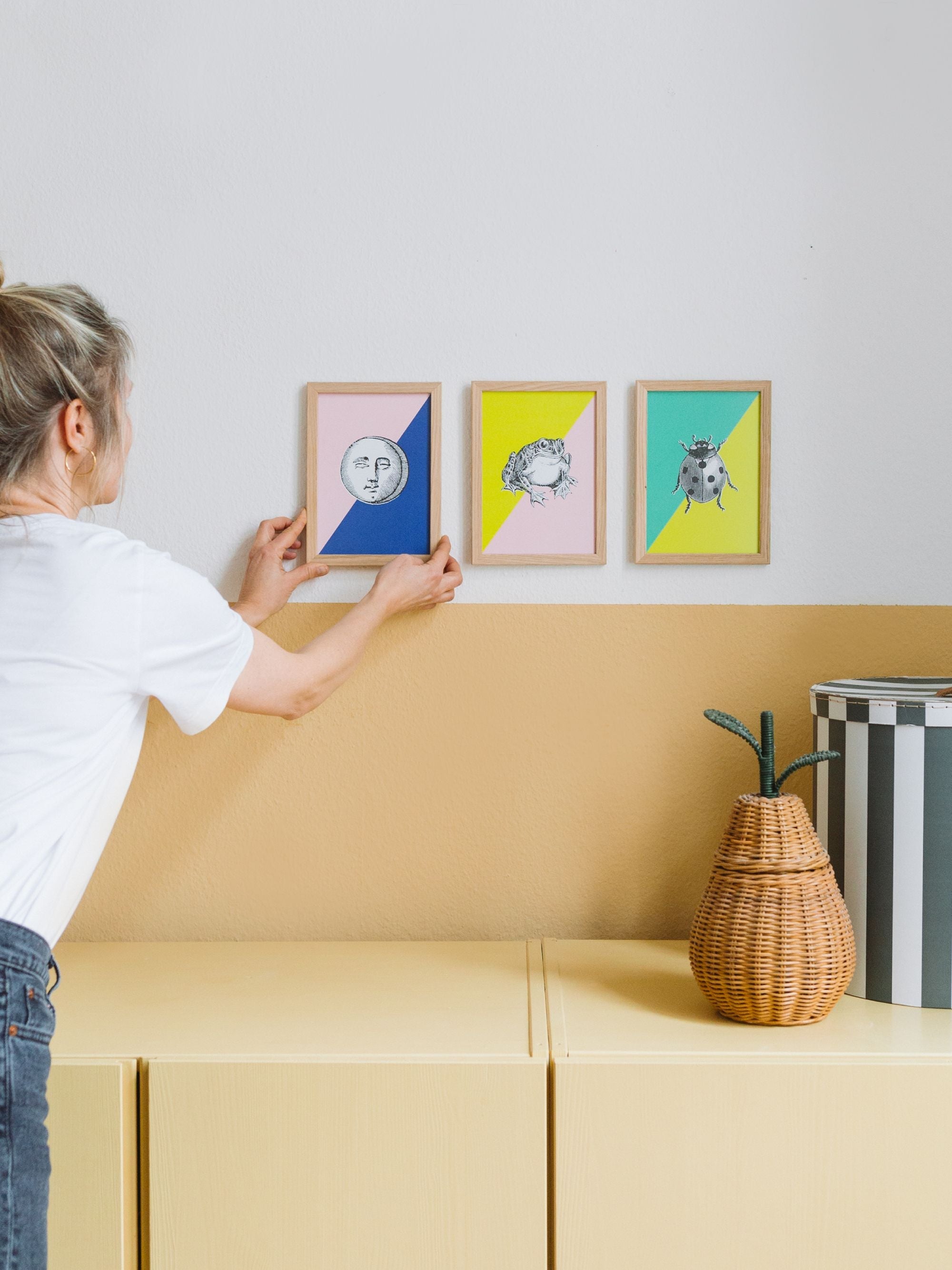 Frau arrondiert kleine gerahmte Kunstdrucke mit Mondgesicht, Frosch und Marienkäfer an weiß-gelber Wand über gelbem Schrank mit Birnenkorb.