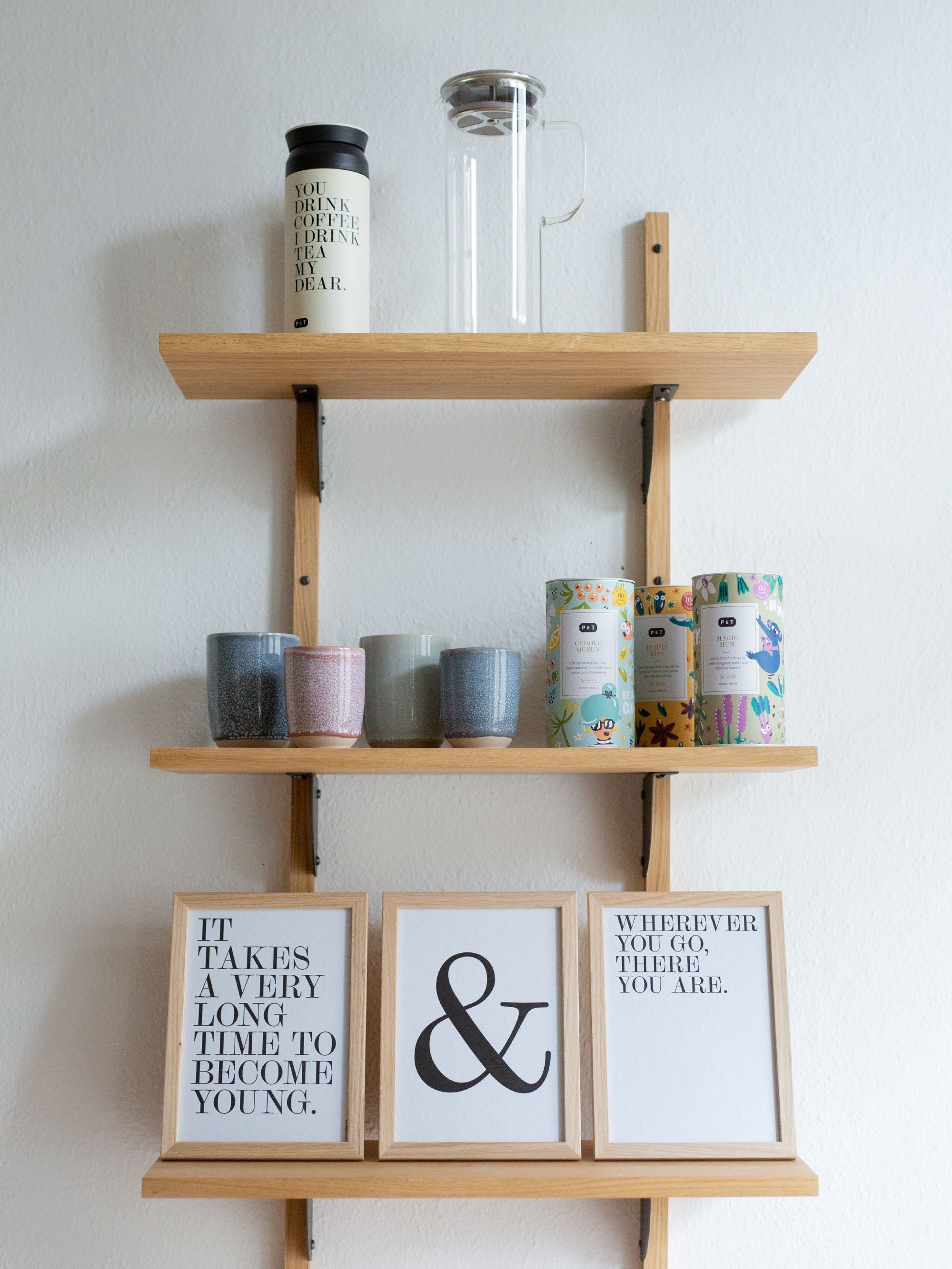 Minimalistisches Holzregal mit weißem Teebecher, transparentem French Press, bunten Keramikbechern und dekorativen Teedosen.