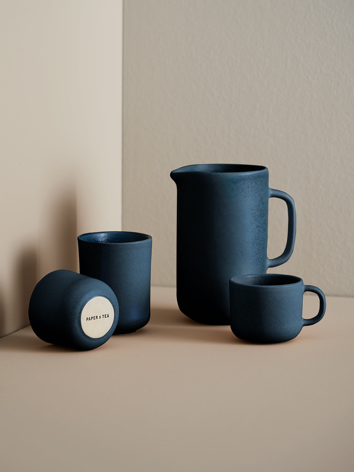 Minimalistisches Set aus mattschwarzer Keramik mit großem Krug und drei Tassen, eine Tasse zeigt PAPER & TEA-Logo.