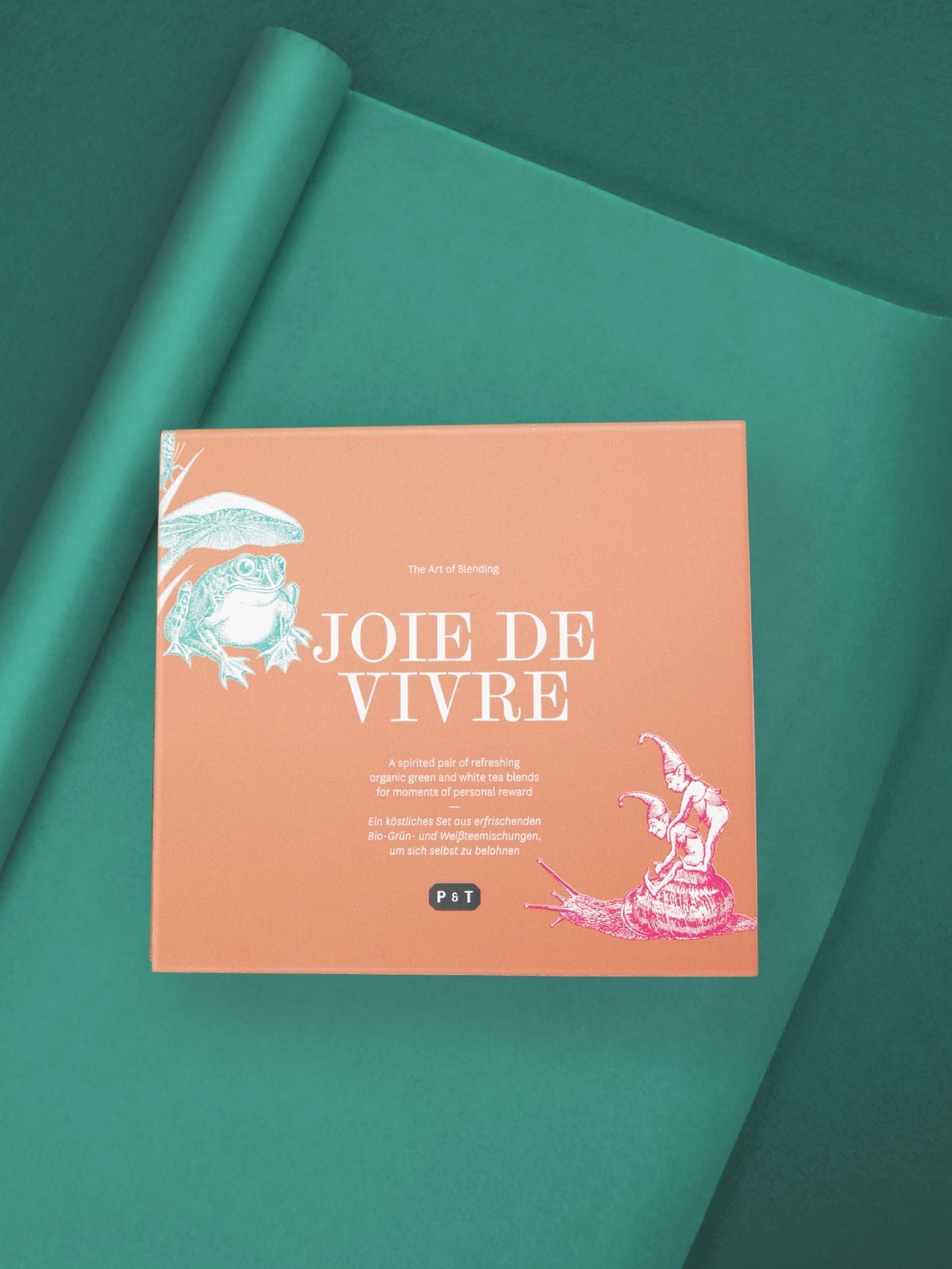 Orangefarbene Box mit weißem Text 'JOIE DE VIVRE Bio-Grün- und Weißtees', auf grünem Hintergrund.