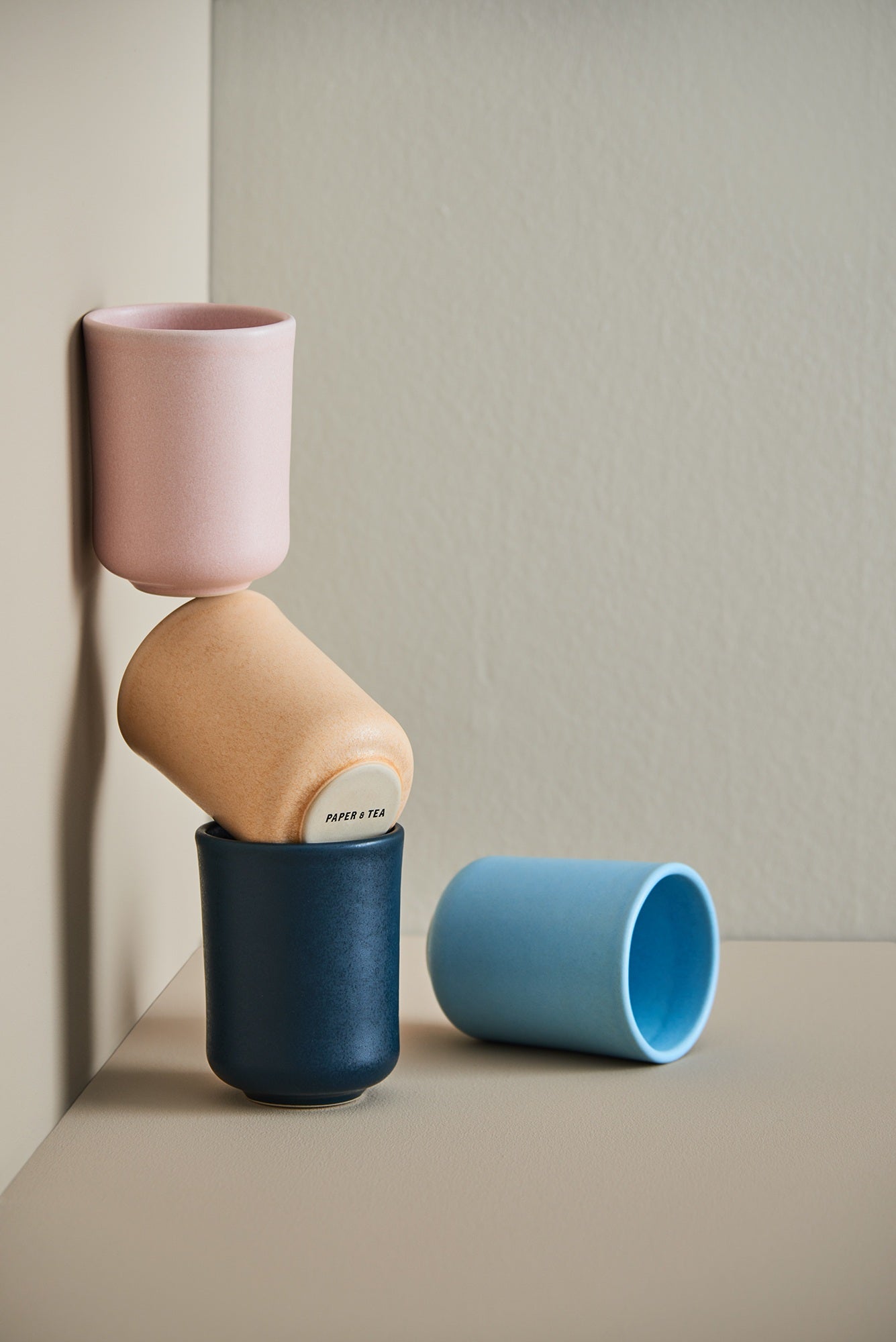Vier minimalistisch gestapelte Keramiktassen in Rosa, Hellbraun mit PAPER & TEA Logo, Dunkelblau und Hellblau vor beiger Wand.
