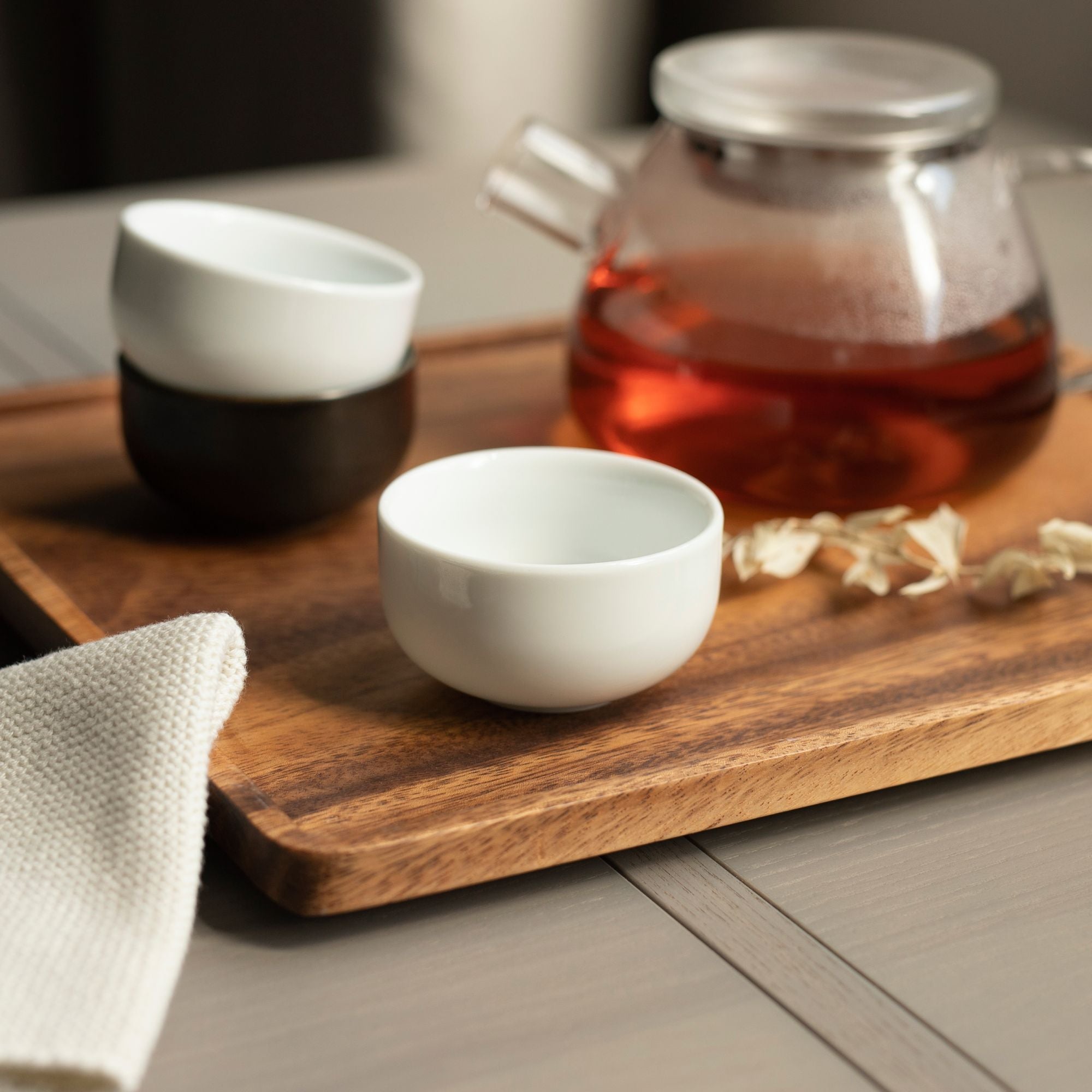Glasteekanne mit metallischem Deckel und rotem Tee auf Holztablett, umgeben von Keramiktassen und Blütenblättern.