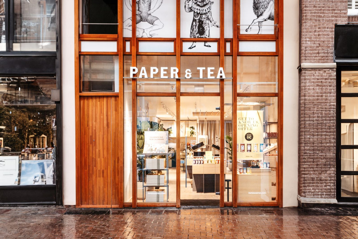Moderne Ladenfront von PAPER & TEA mit Holzrahmen und großen, beleuchteten Fenstern. Werbung zu neuen Kalendern.