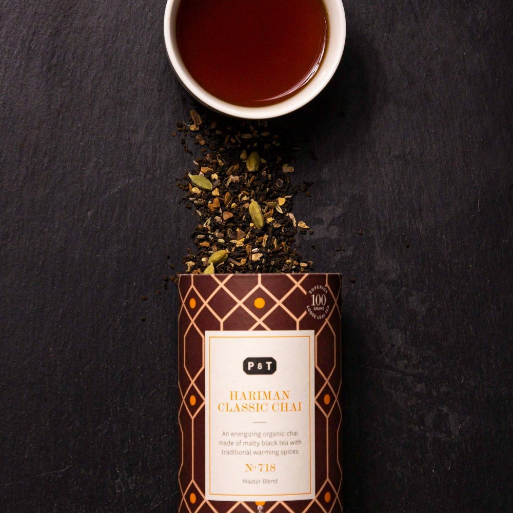 Dose mit HARIMAN CLASSIC CHAI, malziger schwarzer Tee, daneben weiße Tasse Chai und Gewürze auf dunklem Hintergrund.