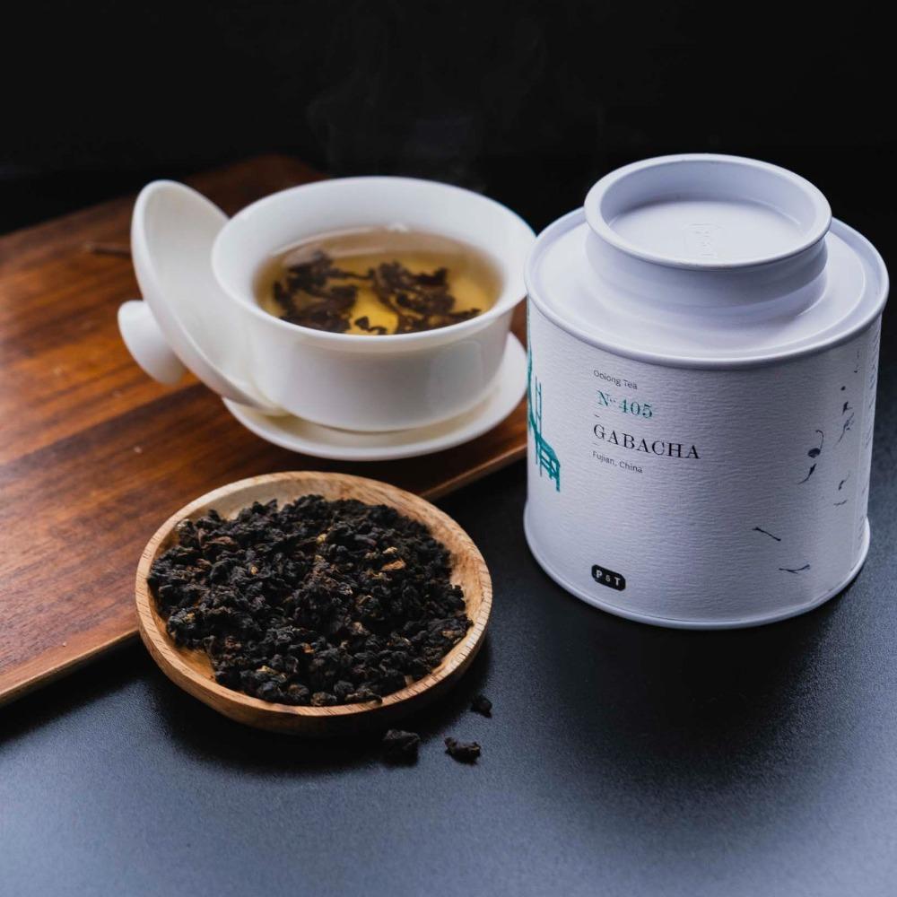 Weiße Zylinderdose mit Oolong Tee Gabacha N-405 aus Fupan, daneben Tasse Bernstein-Tee und Tablett mit Teeblättern.