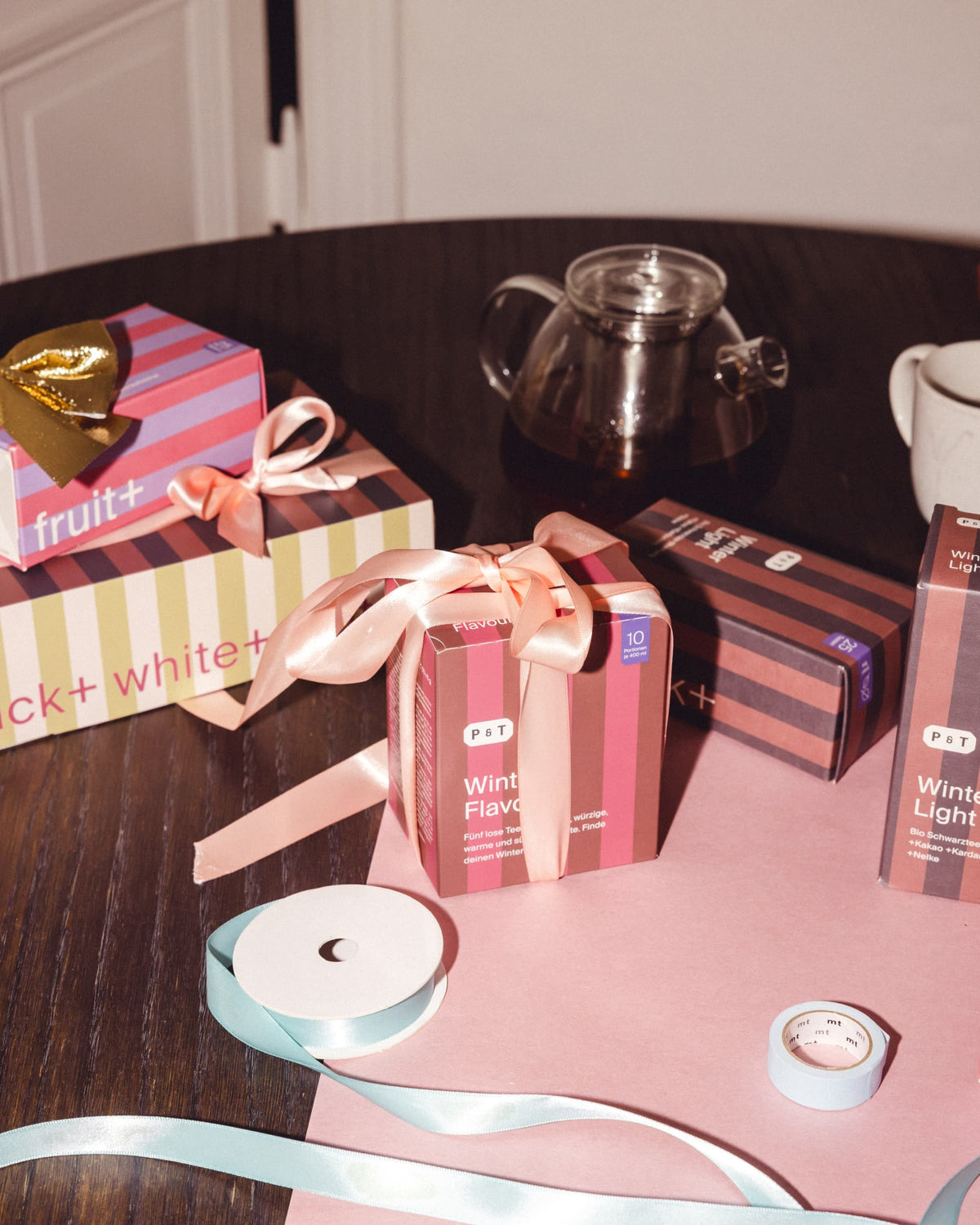 Vier gestreifte P&T Geschenkboxen mit rosa Schleifen und Glas-Teekanne auf Holztisch, winterliche Teemischungen.