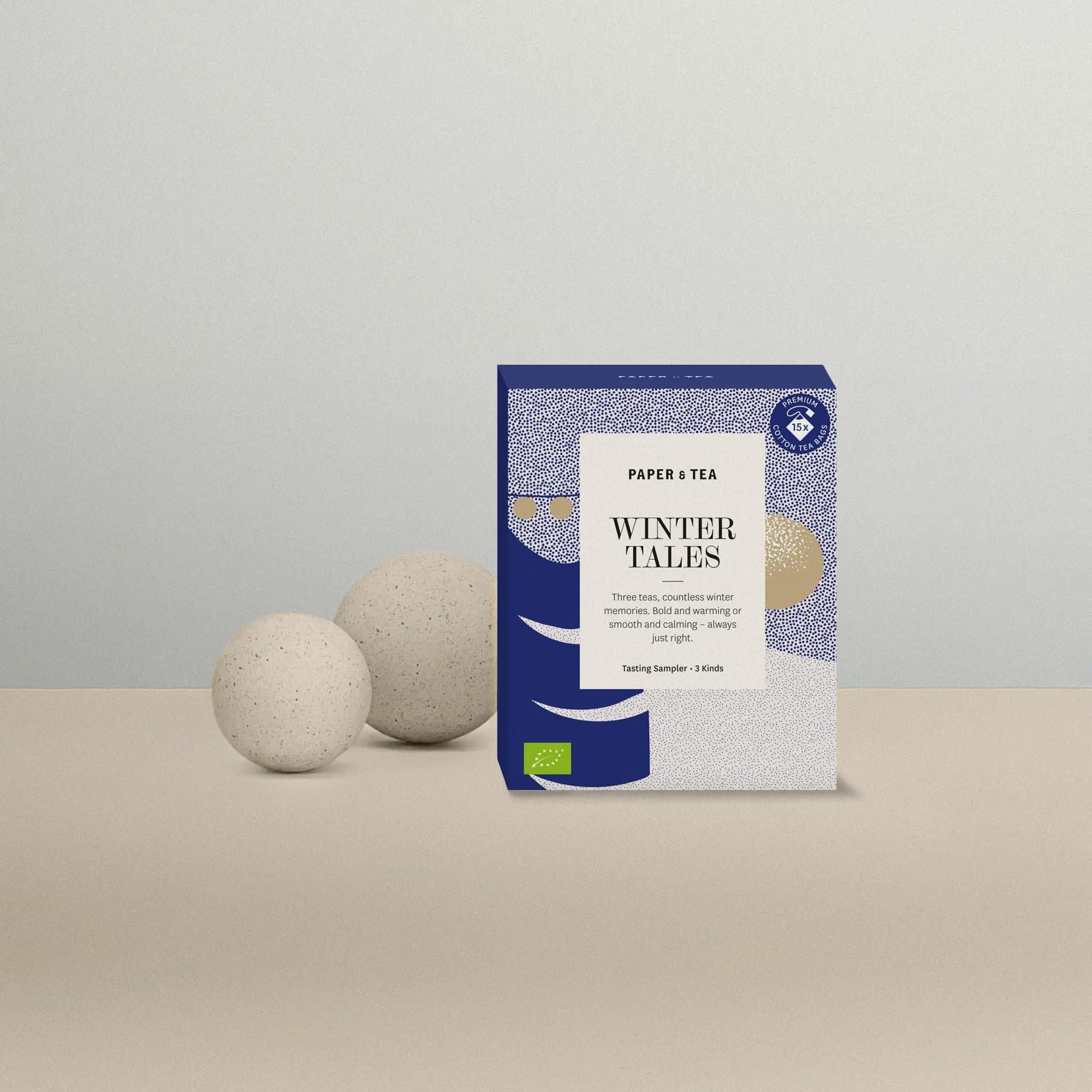 PAPER & TEA WINTER TALES Probierpaket mit drei Tees in blauer und beiger Verpackung neben Deko-Kugeln.