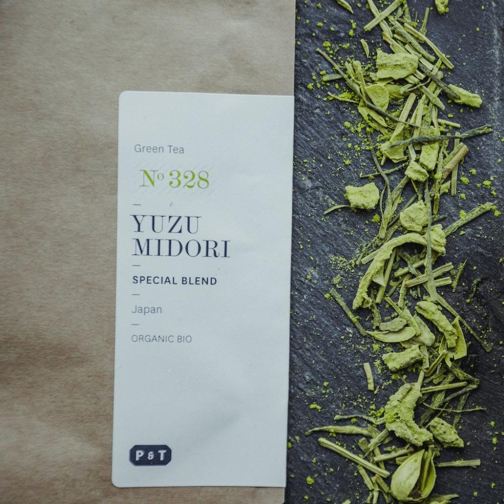 Weißes Etikett mit der Aufschrift YUZU MIDORI, Bio-Grüntee aus Japan, neben losen Teeblättern auf dunklem Hintergrund